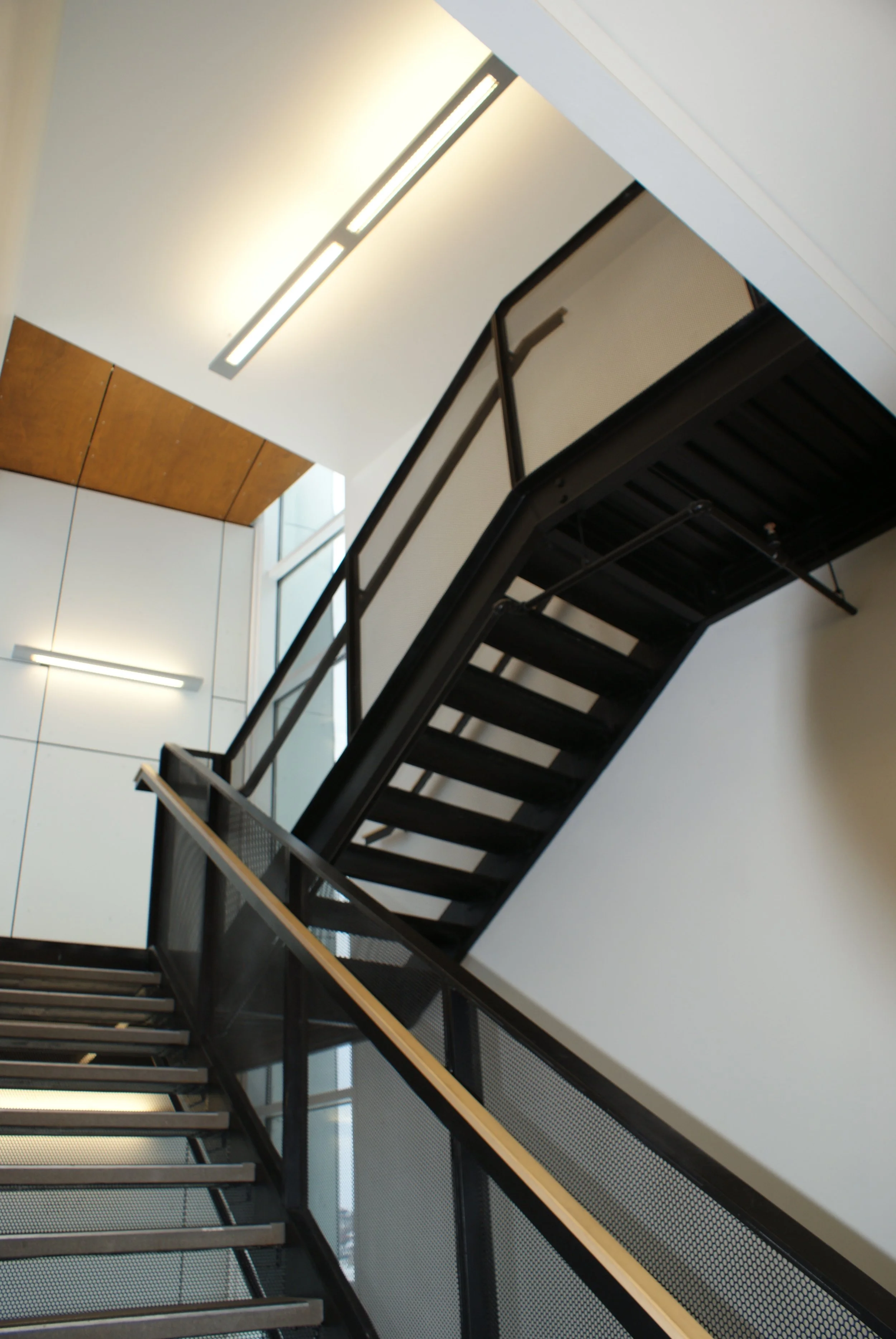 Interior view of a modern stairwell with black metal stairs and railings, white walls, ceiling lights, and a wooden panel on the ceiling.