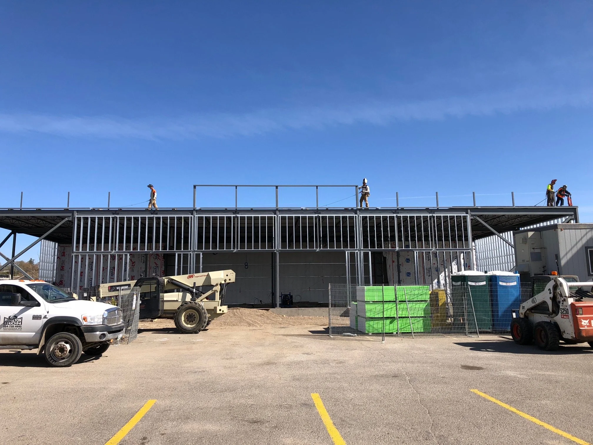 Construction workers on the roof of a building under construction, with construction equipment and supplies in the foreground, and a clear blue sky.