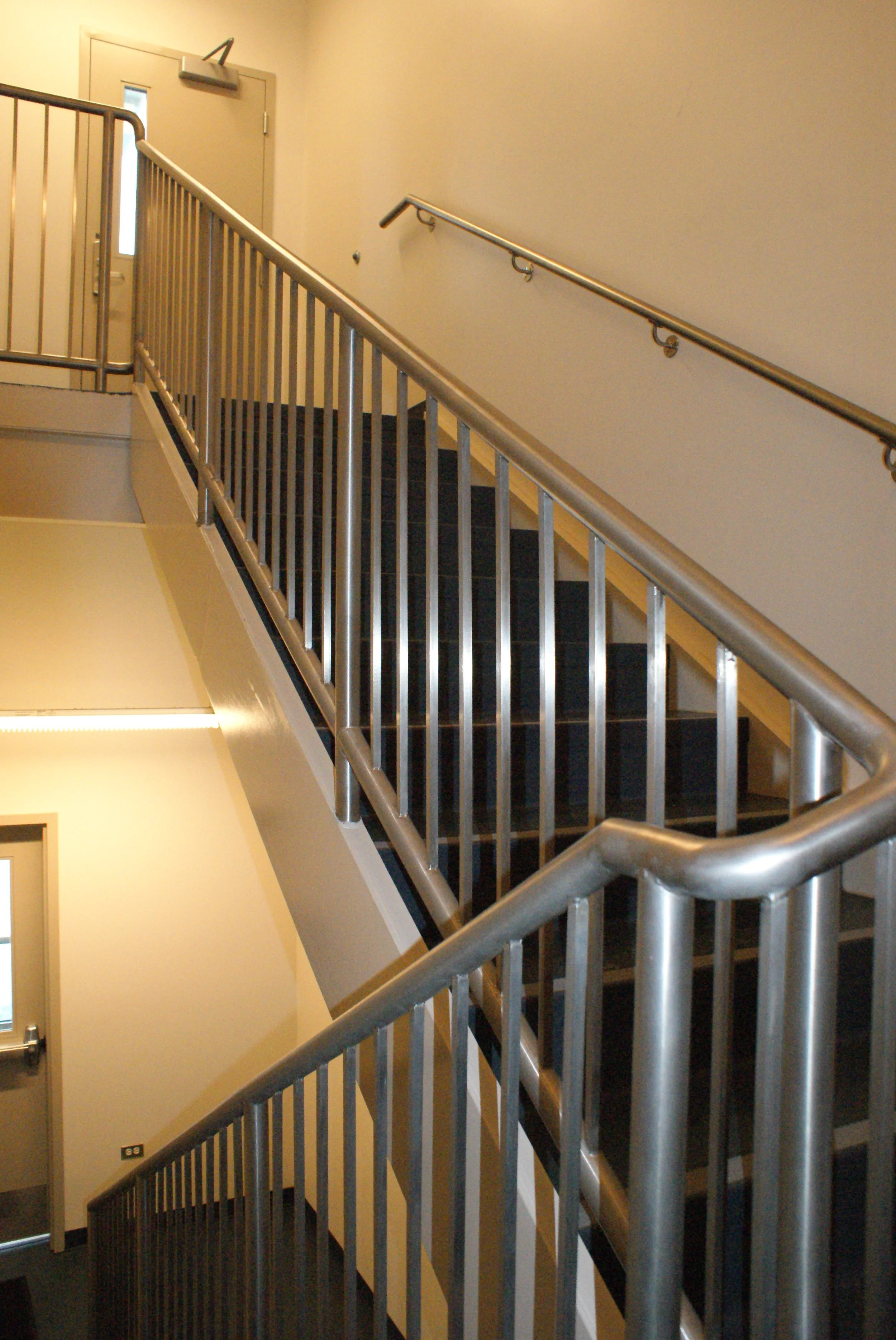 An indoor staircase with metal handrails and black steps, leading to a door at the top, in a beige-walled building.