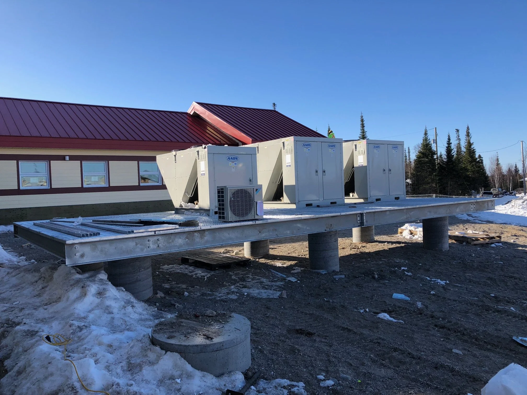 Construction site with a metal platform supported by concrete piers, HVAC units, and a building with a red metal roof and yellow siding in the background, snowy ground, and a clear blue sky.
