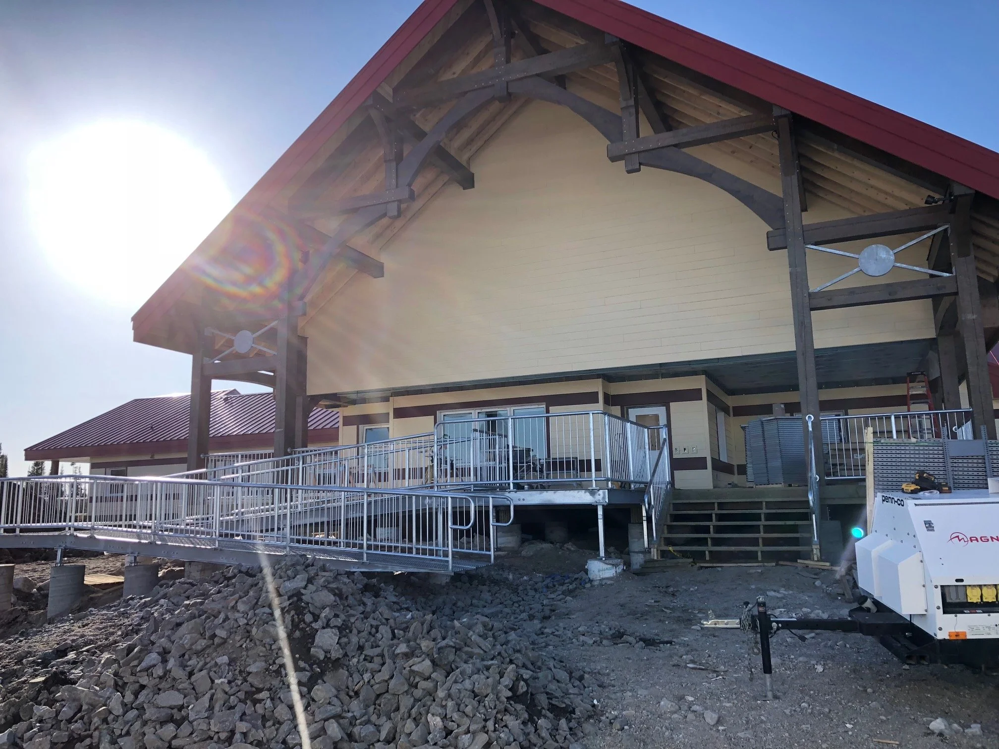 A large house under construction with an unfinished foreground of rocks and dirt, a metal ramp on the left side, and construction equipment on the right, with the sun shining brightly in the sky.