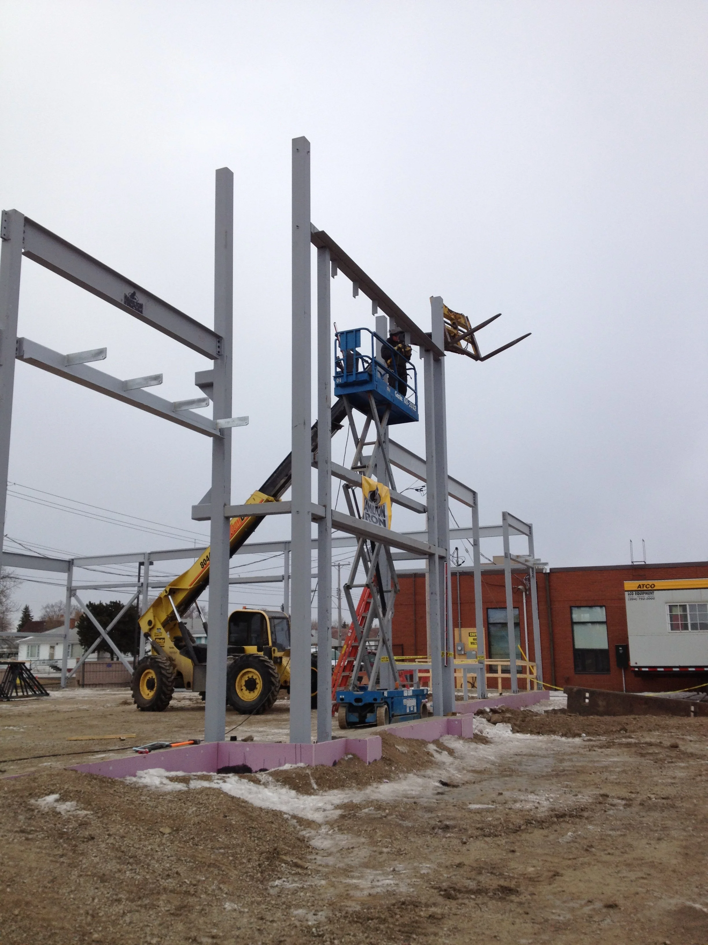 Construction site with steel framework, a yellow forklift, and a worker on a blue lift working on erecting the structure.