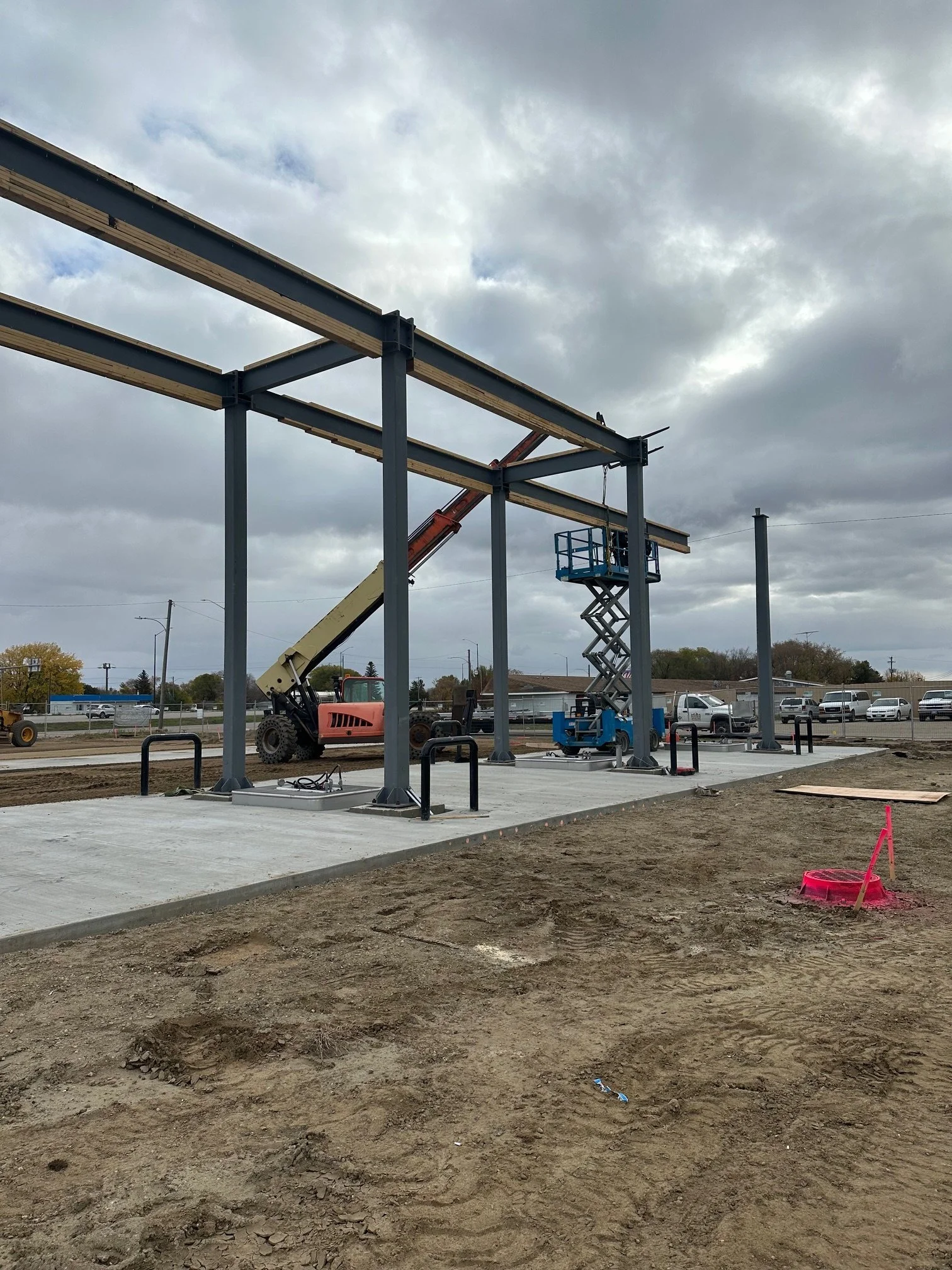 Construction site with steel framework and a crane, with cloudy sky and parked cars in the background.