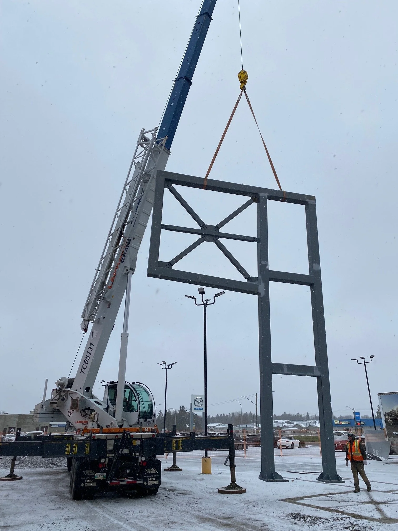 A crane lifting a large metal framework structure in a snowy parking lot, with workers supervising and a Mazda dealership sign in the background.