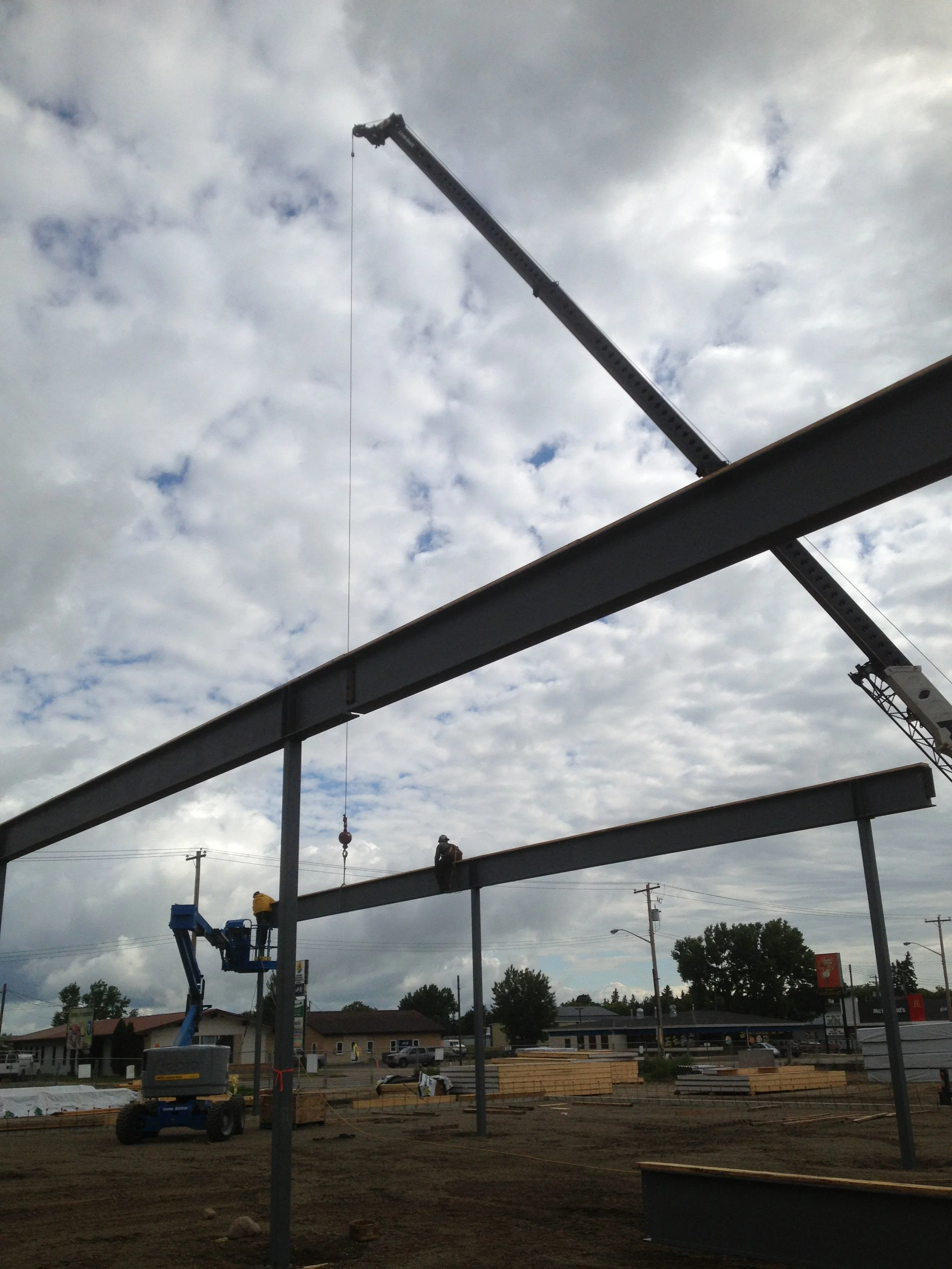 Construction site with steel beams being assembled, workers on the beams, a crane lifting a beam, and a cloudy sky overhead.