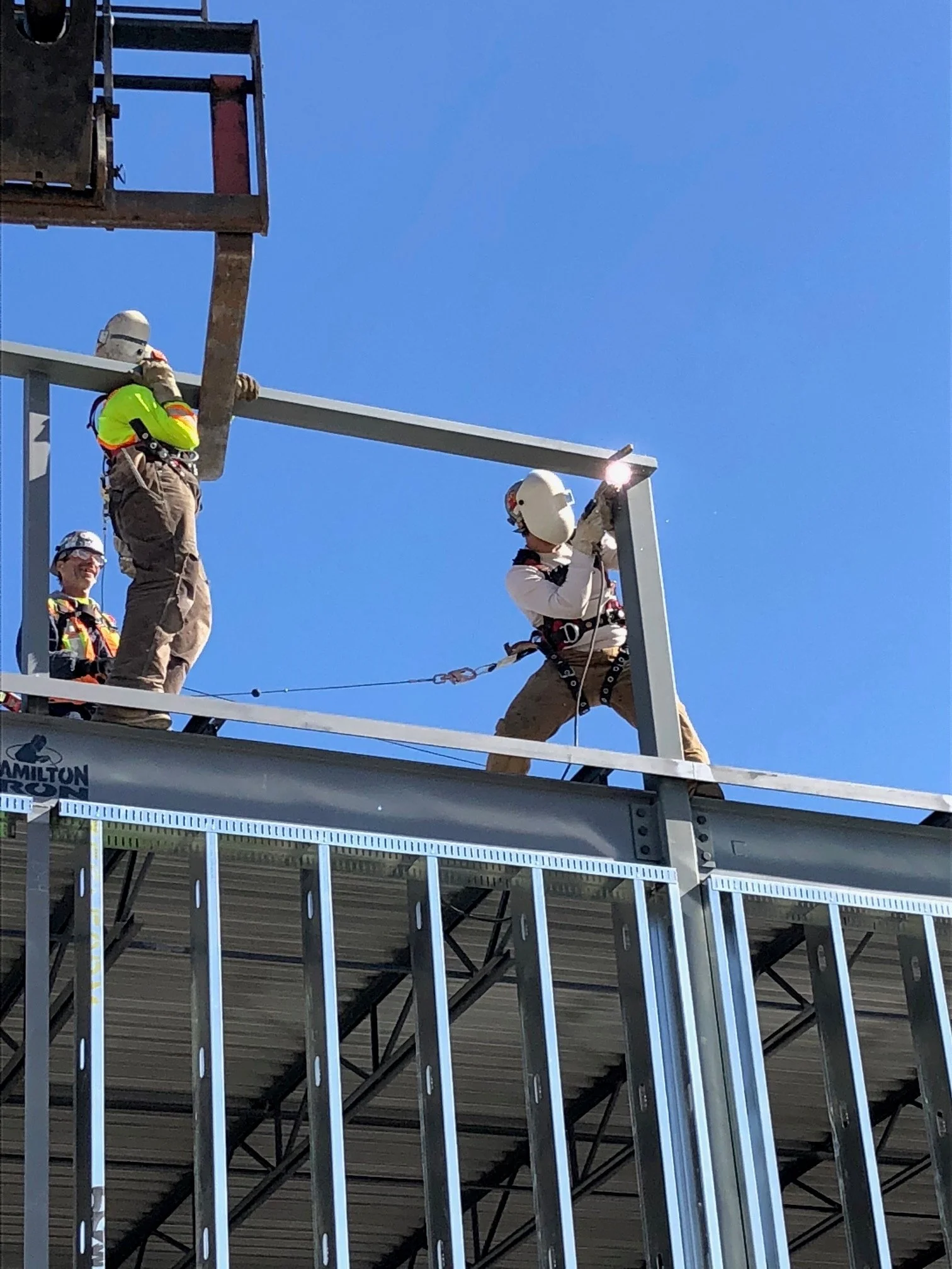 Construction workers with safety gear installing a structural beam on a building under construction against a clear blue sky.