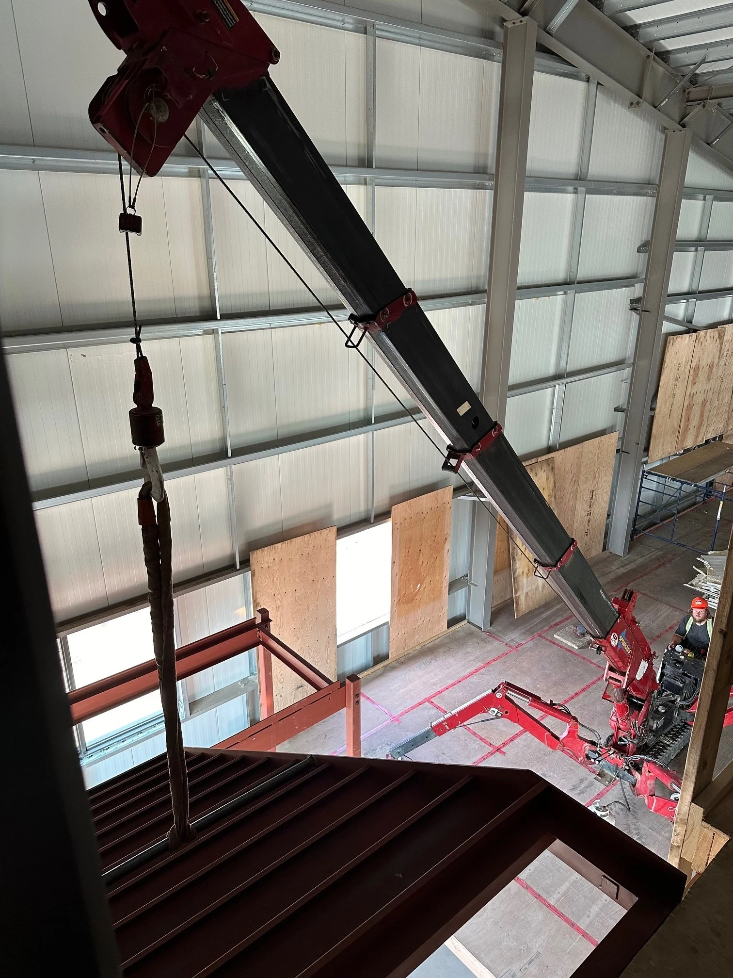 Inside a warehouse with a large red crane being operated by a worker wearing a helmet, lifting a heavy object.