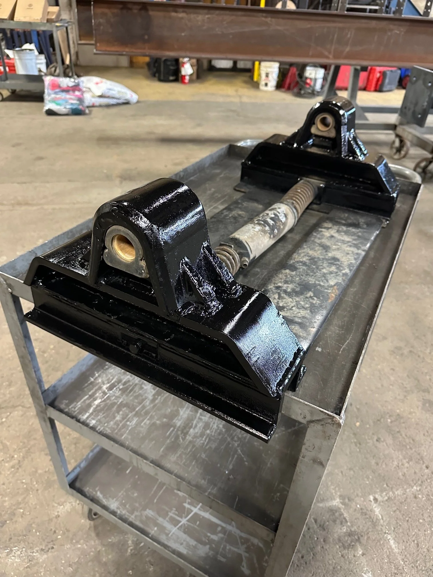 A metal car suspension component with black painted parts on a stainless steel worktable in a workshop.