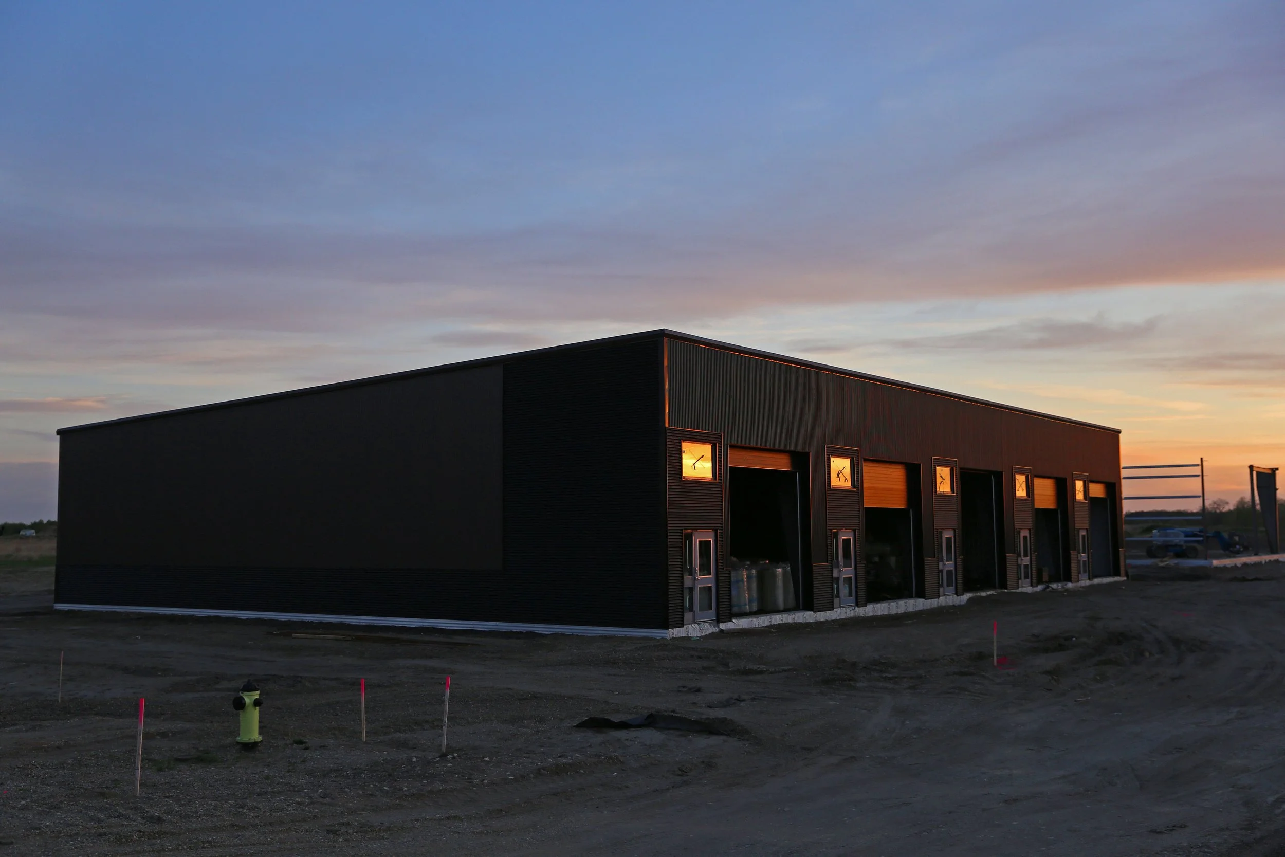 A modern industrial building with black exterior walls, large open garage doors, and small windows with lamps inside, set against a sunset sky and a construction area with equipment and dirt ground.