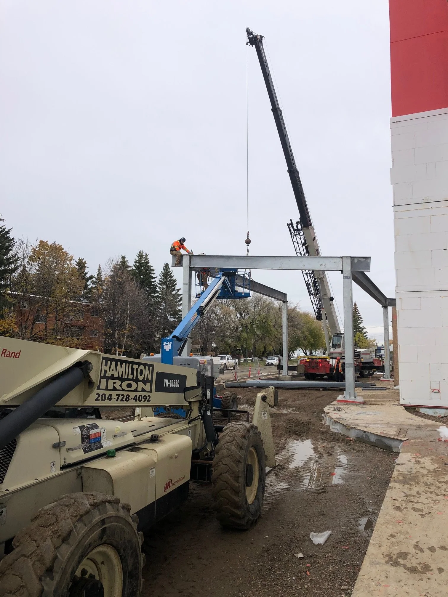 Construction workers are using cranes and lifts to assemble steel framework on a building site, with equipment and muddy ground in the foreground.
