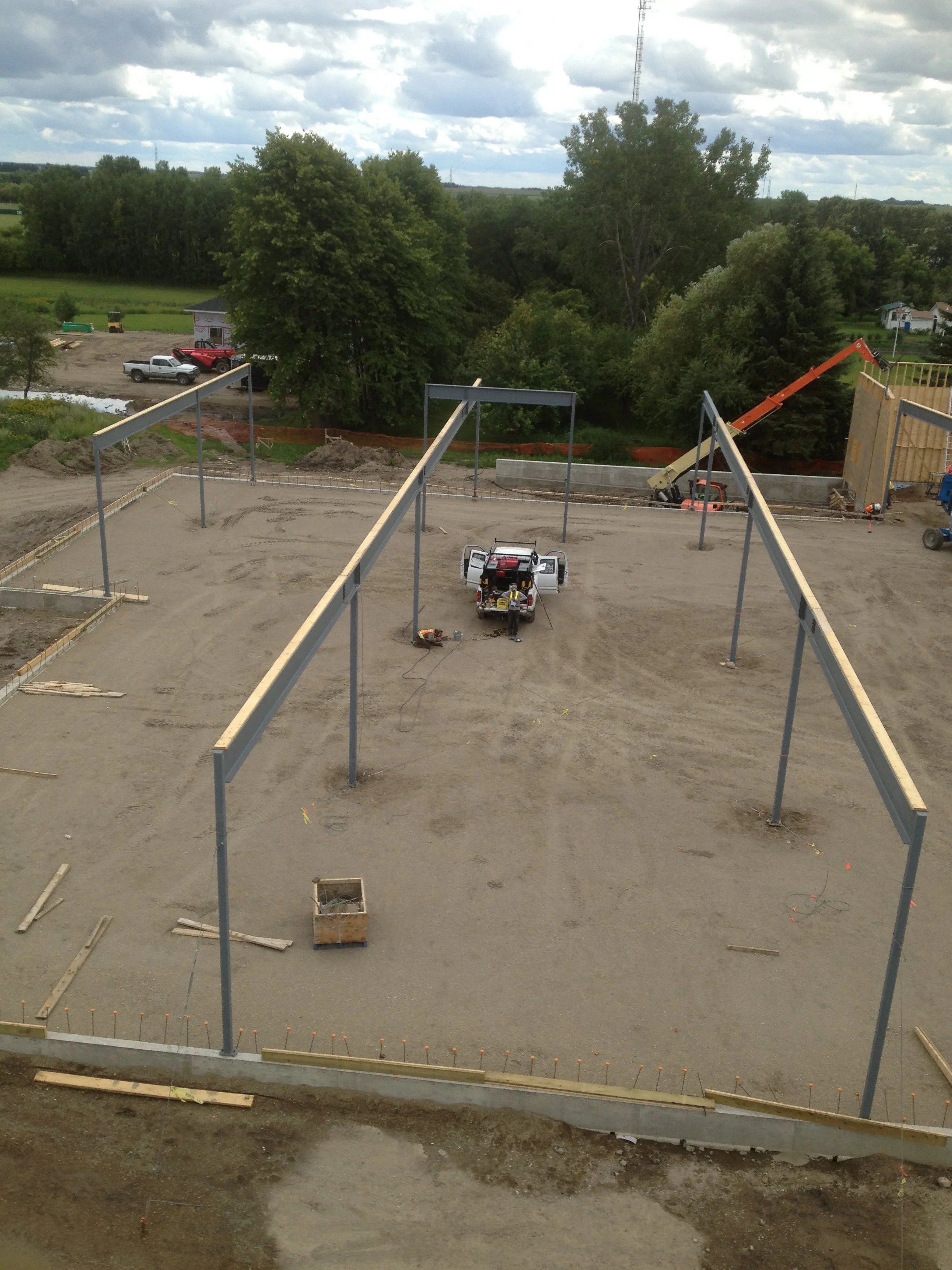 Construction site with steel framing for a building, a small vehicle, and construction equipment on dirt ground. Green trees and a cloudy sky in the background.