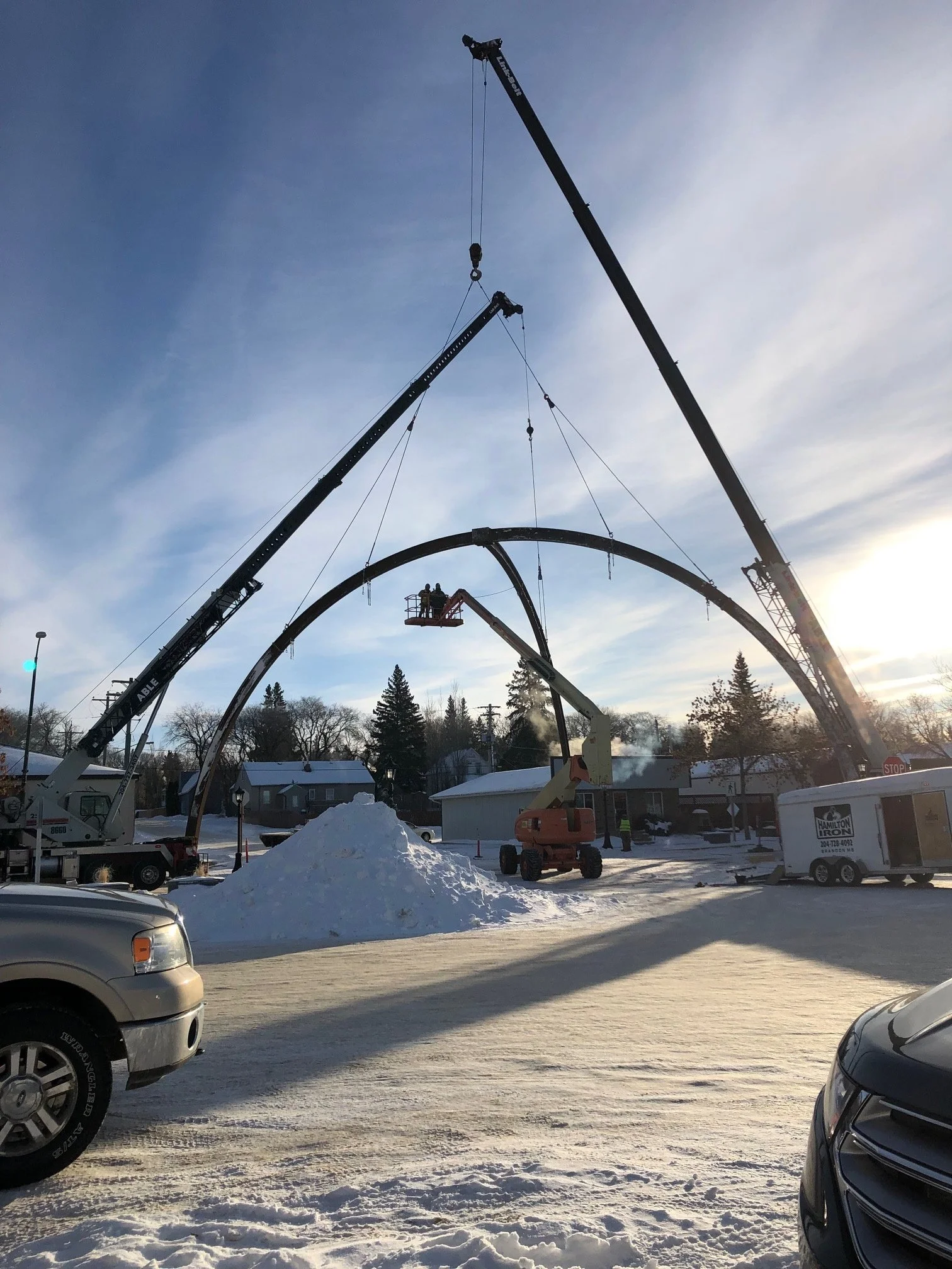 Construction workers on a cherry picker installing a large arch structure in a snowy urban area with cranes and vehicles.