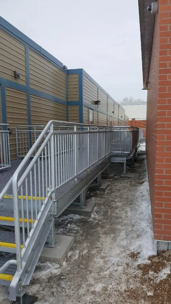 Metal wheelchair accessible ramp with handrails next to a brick building, with snow and ice on the ground.