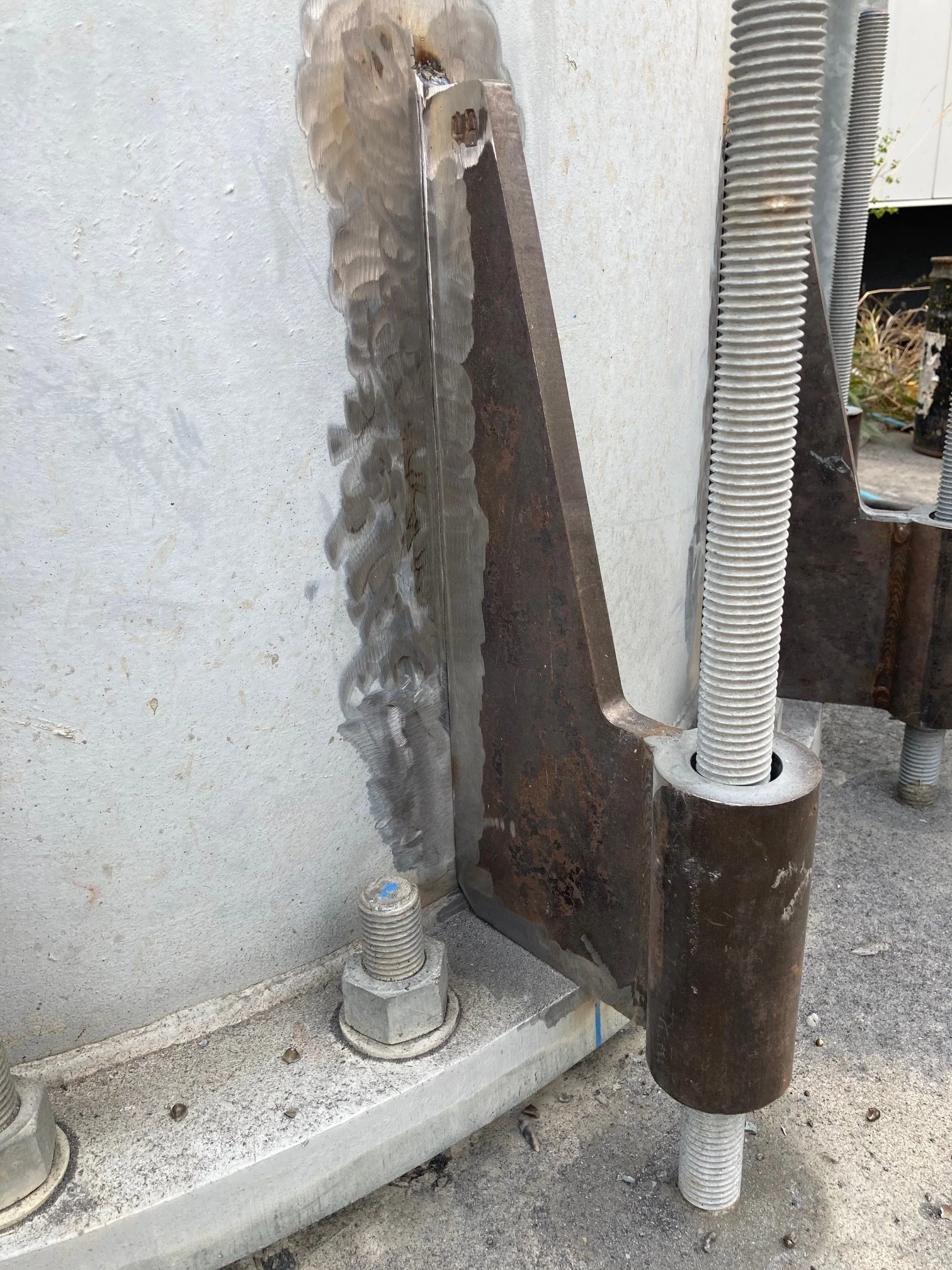 Close-up of a large metal anchor bolt and steel plate attached to a concrete surface, with a rusted metal support structure and flexible conduit pipe nearby.
