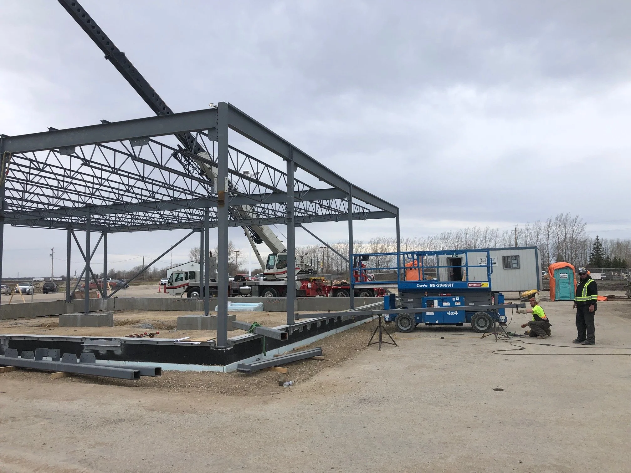 Construction workers building a metal structure with a crane, blue lift, and portable toilet on site under a cloudy sky.