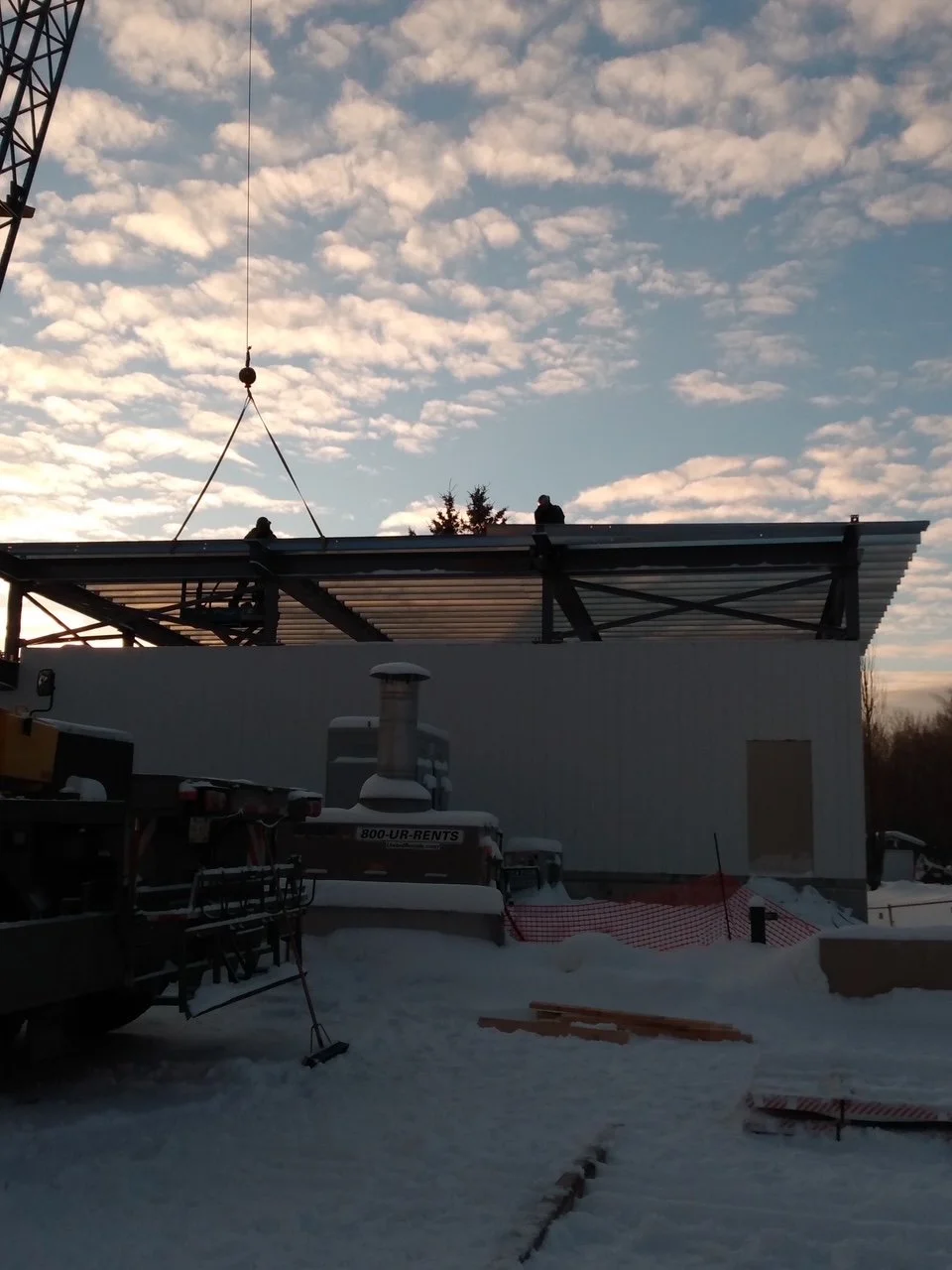 Construction workers installing a metal roof on a building during winter, with snow on the ground and a sky filled with scattered clouds.