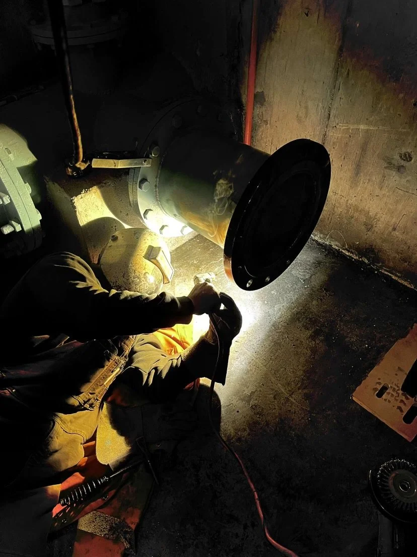 Worker welding a large pipe in a dark industrial setting, wearing a welding helmet and protective clothing.