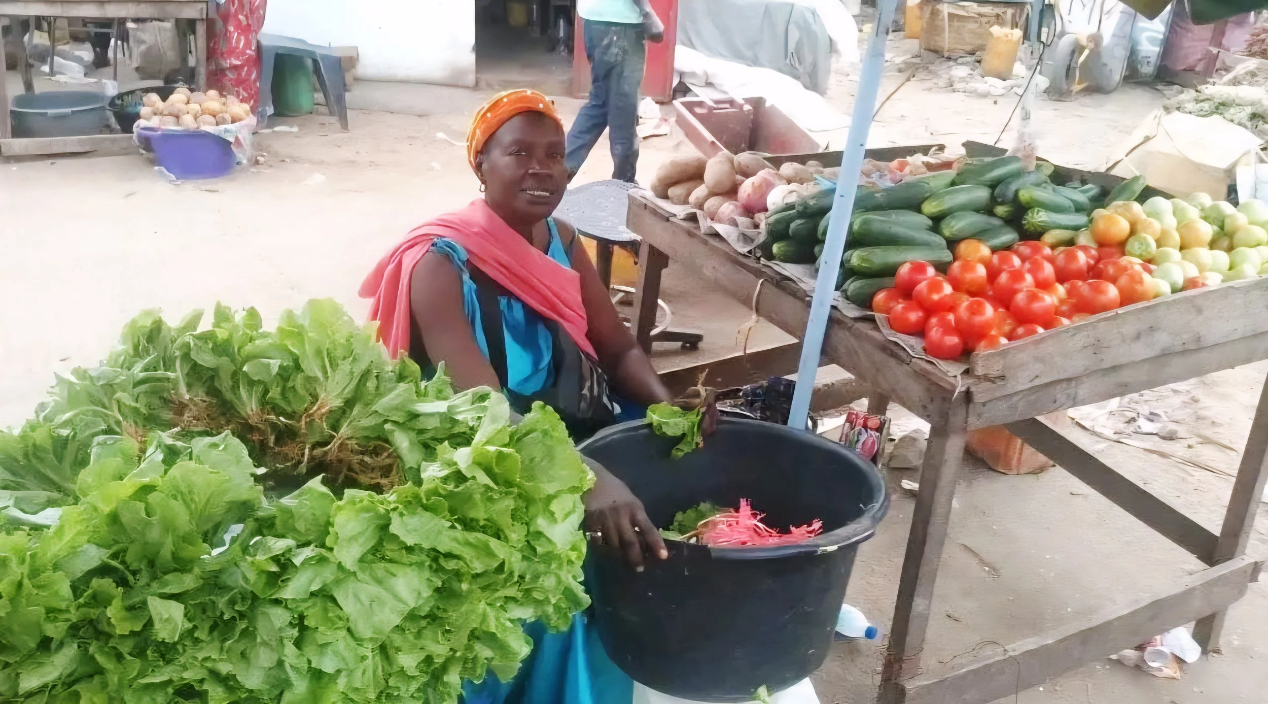 De moeder van Saikou op een lokale groentemarkt in Senegal, omringd door verse sla, tomaten, komkommers en uien.