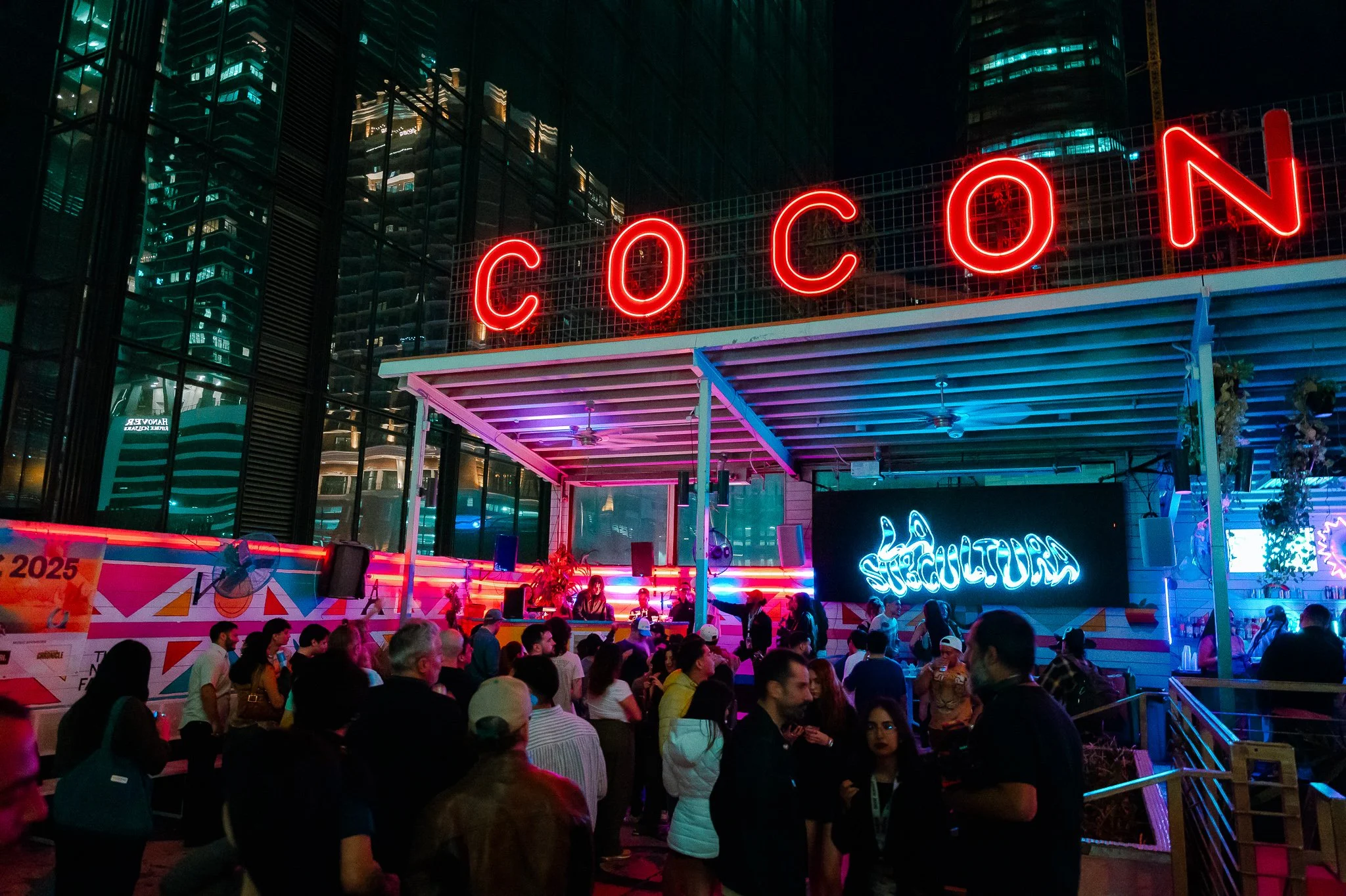 Night scene of an outdoor event at a venue called 'Cocoon' with a large illuminated red sign, a brightly lit stage with DJ equipment, neon lights, and a crowd of people socializing under a large modern glass building.