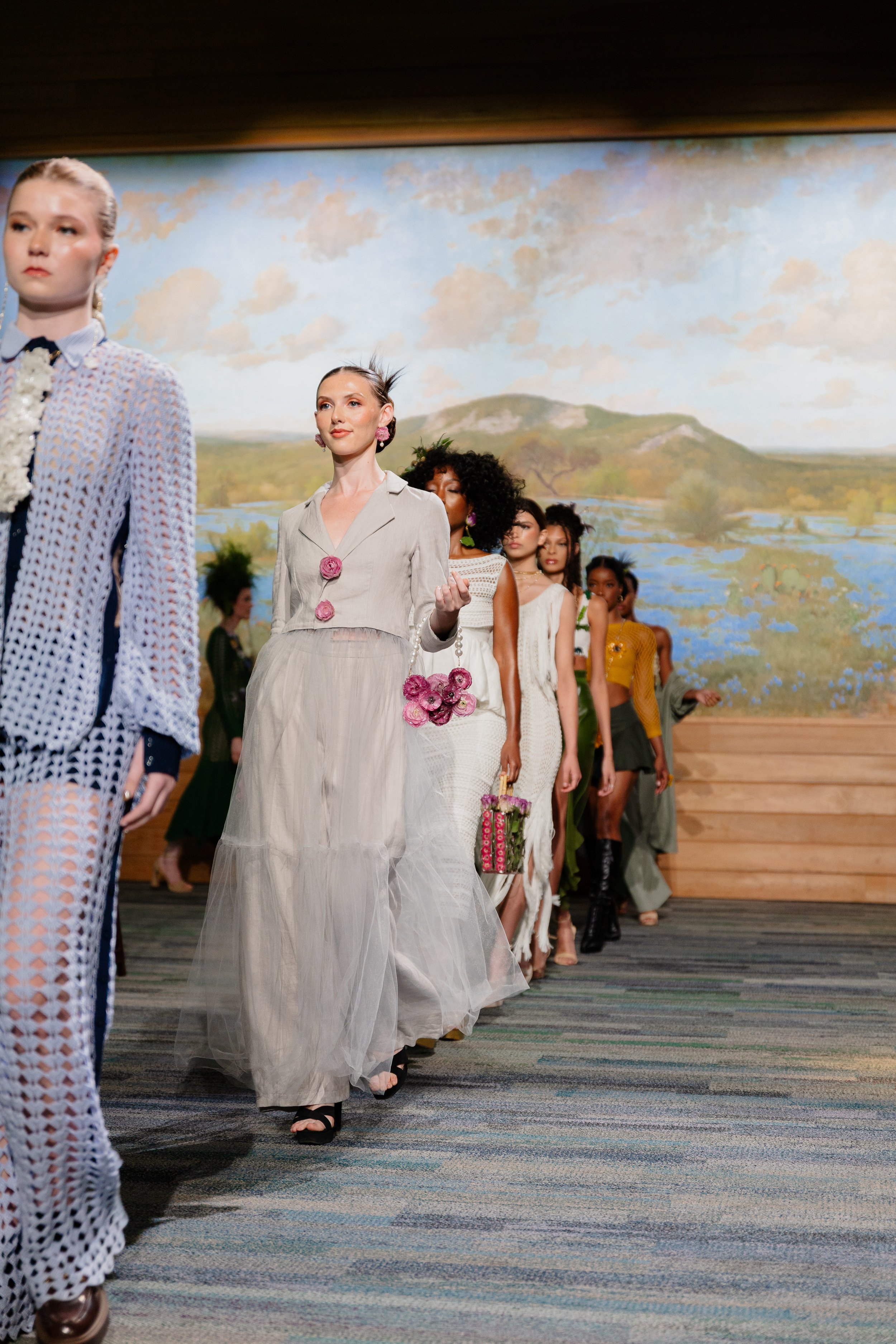 Models walking down a runway during a fashion show, with a painted landscape backdrop.