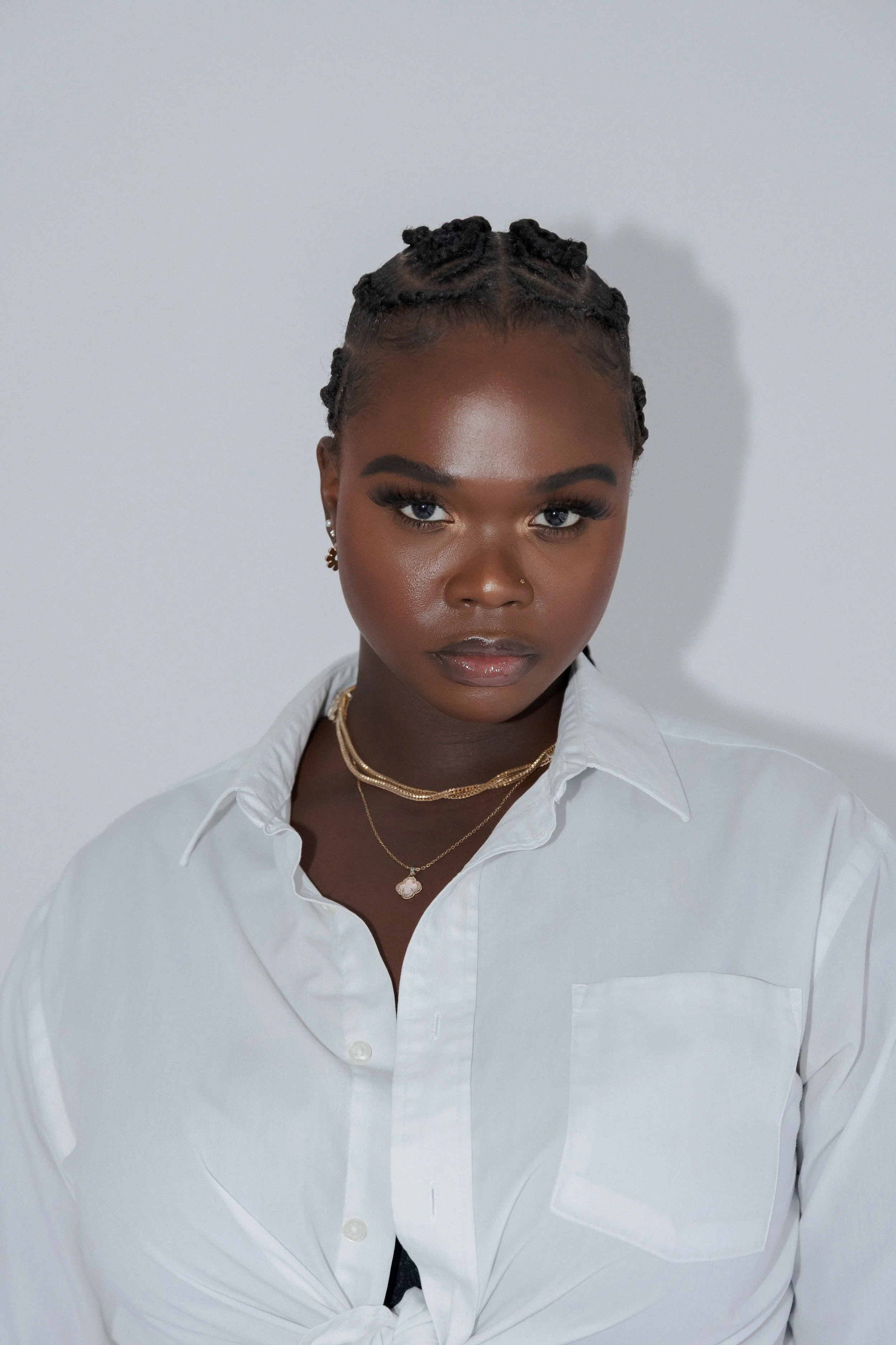 Portrait of a woman with dark skin, styled hair, wearing a white button-up shirt, layered gold necklaces, and earrings, standing against a plain white background.