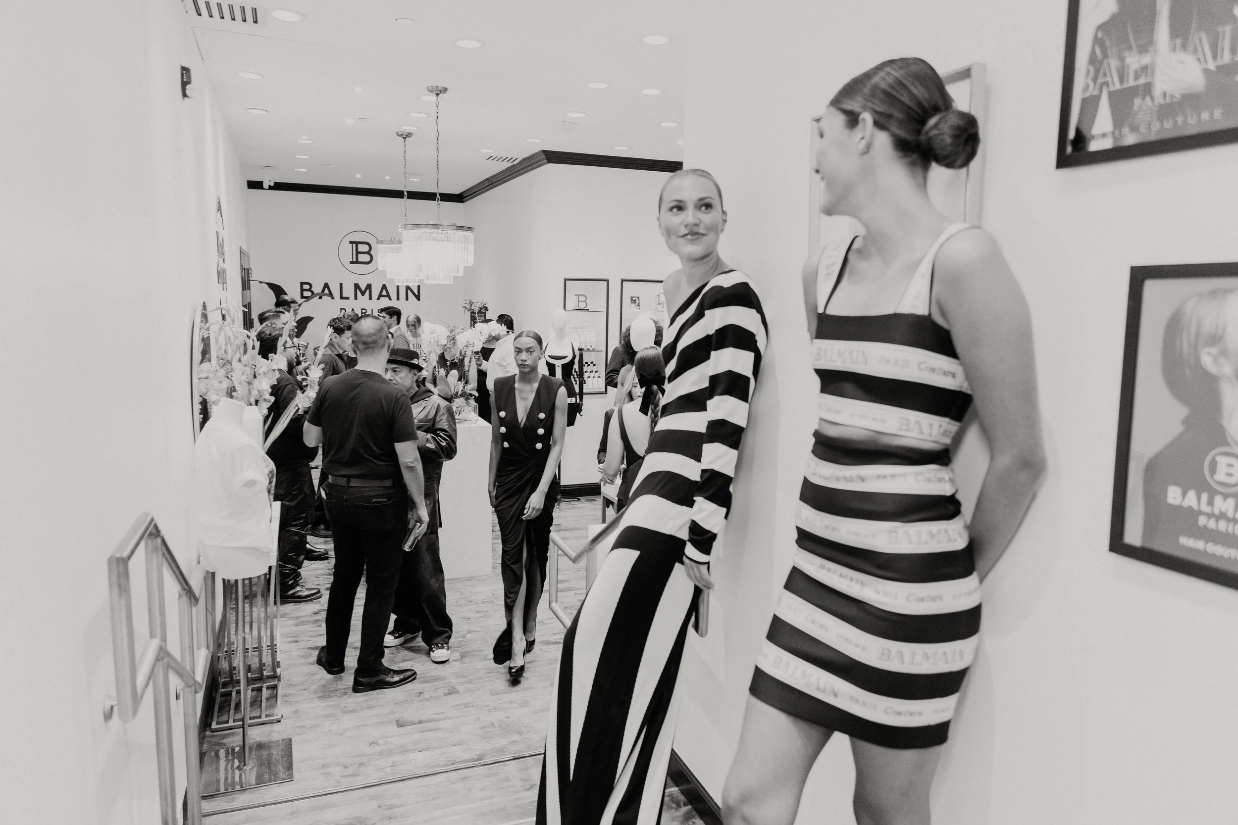 Black and white photo of a fashion store with mannequins in striped dresses and people browsing, featuring the Balmain logo on the back wall.
