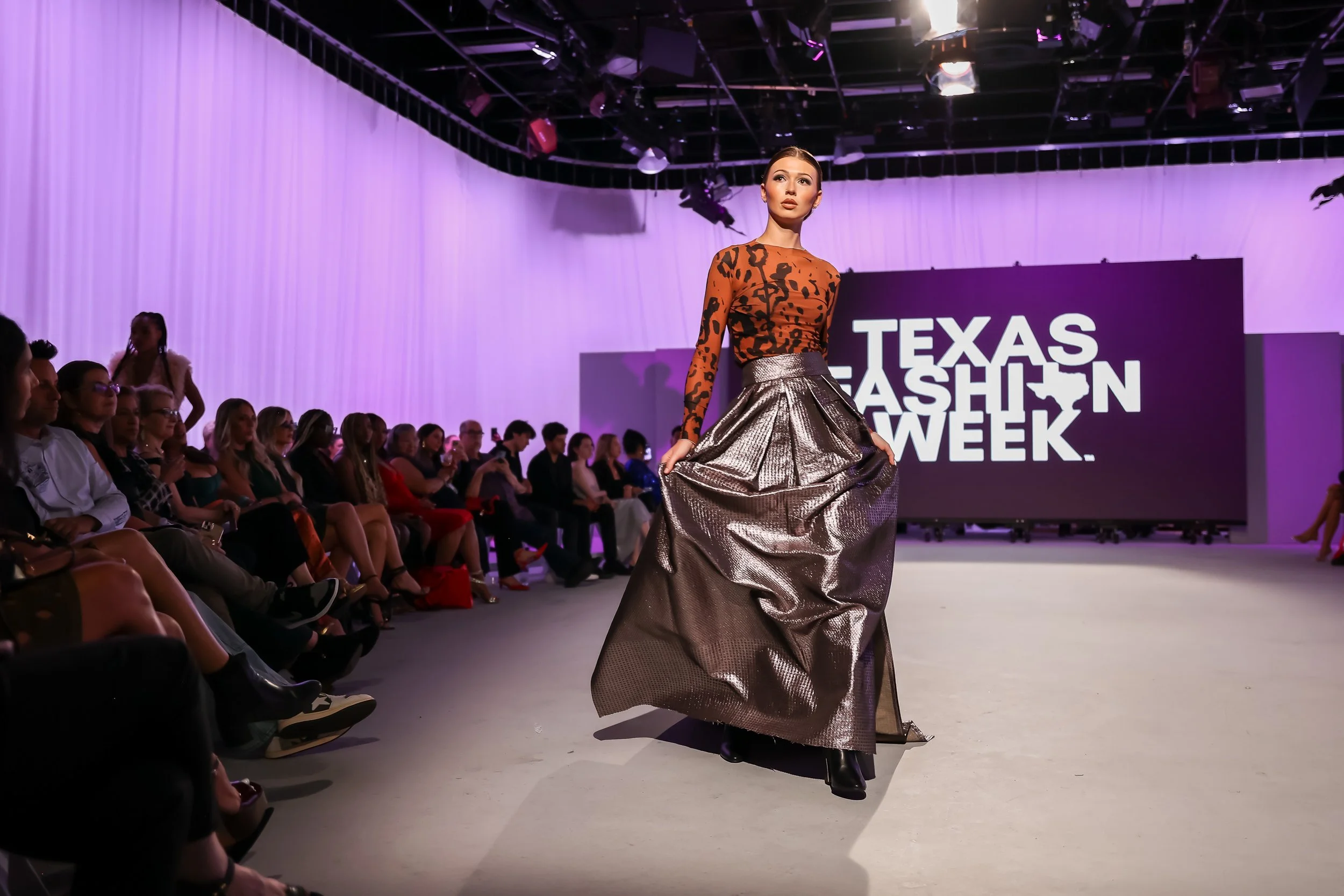 A female model walking on the runway during Texas Fashion Week, wearing a metallic silver high-low skirt and a long-sleeved, animal print top. The audience watches attentively as she poses with a confident expression.