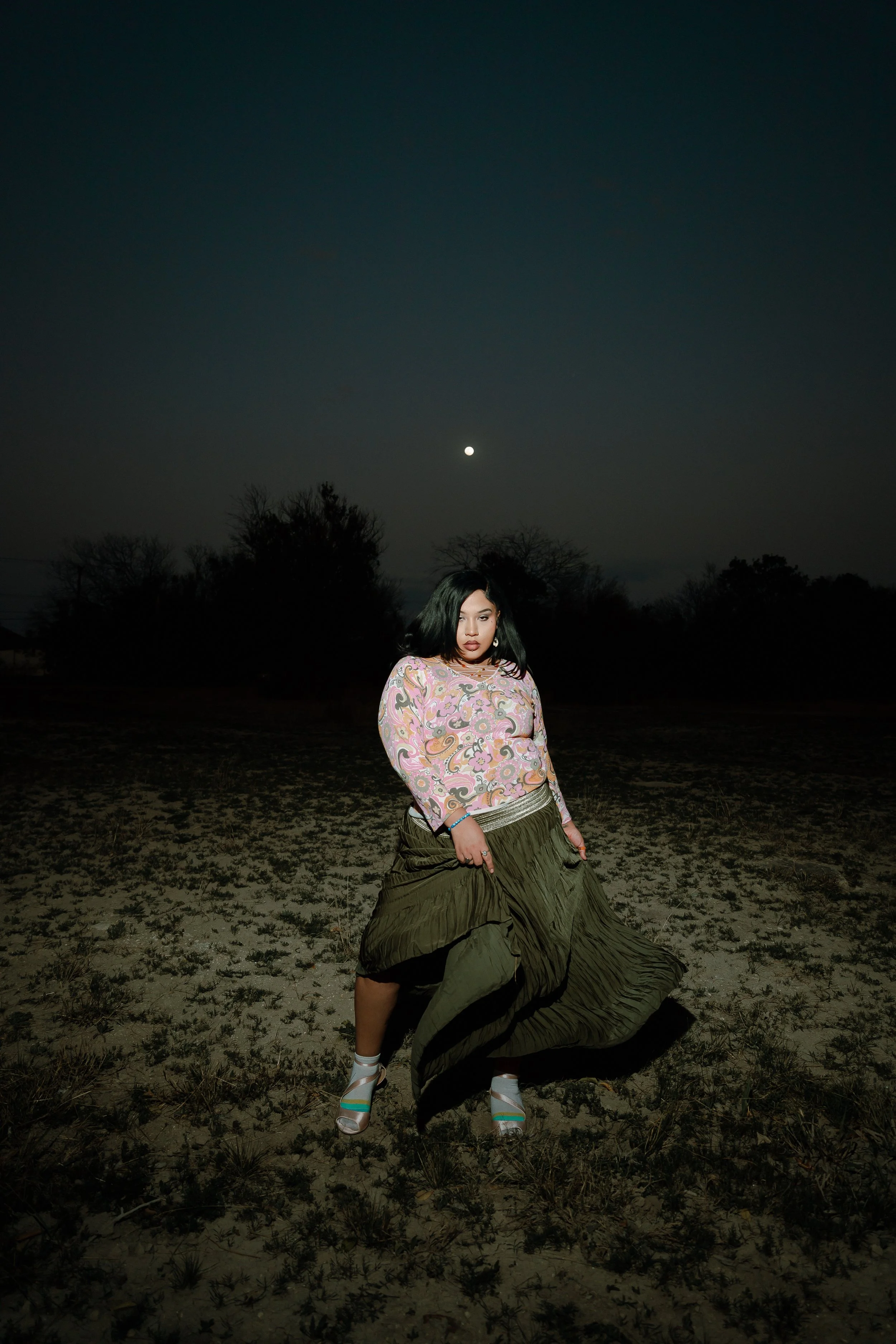 A woman standing outdoors at night, illuminated by artificial light, with a dark sky and a bright moon overhead, wearing a patterned long-sleeve top, loose green skirt, and high-heeled shoes.