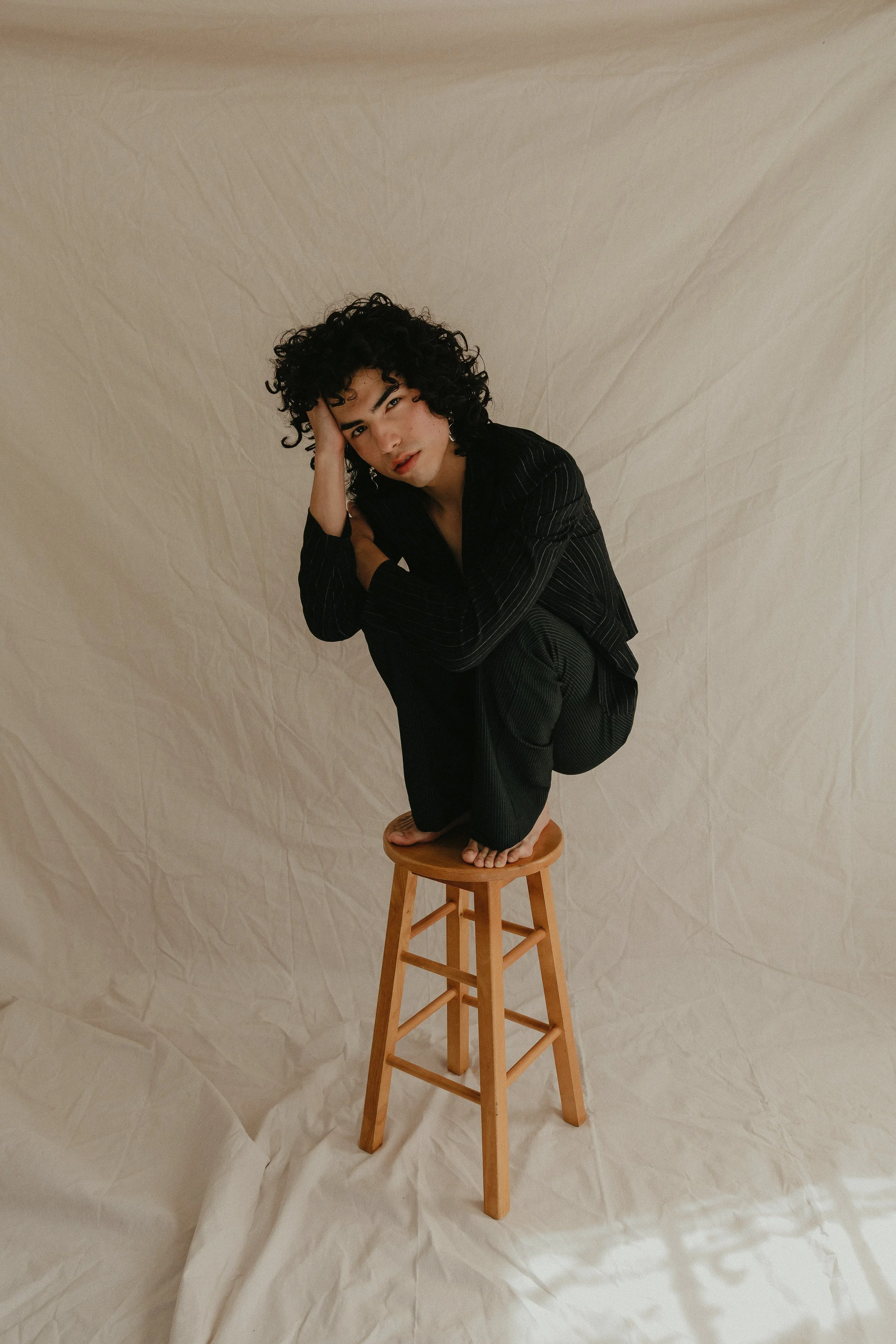 A woman with curly hair dressed in black, squatting on a wooden stool against a beige backdrop, with her head resting on one hand and looking at the camera.