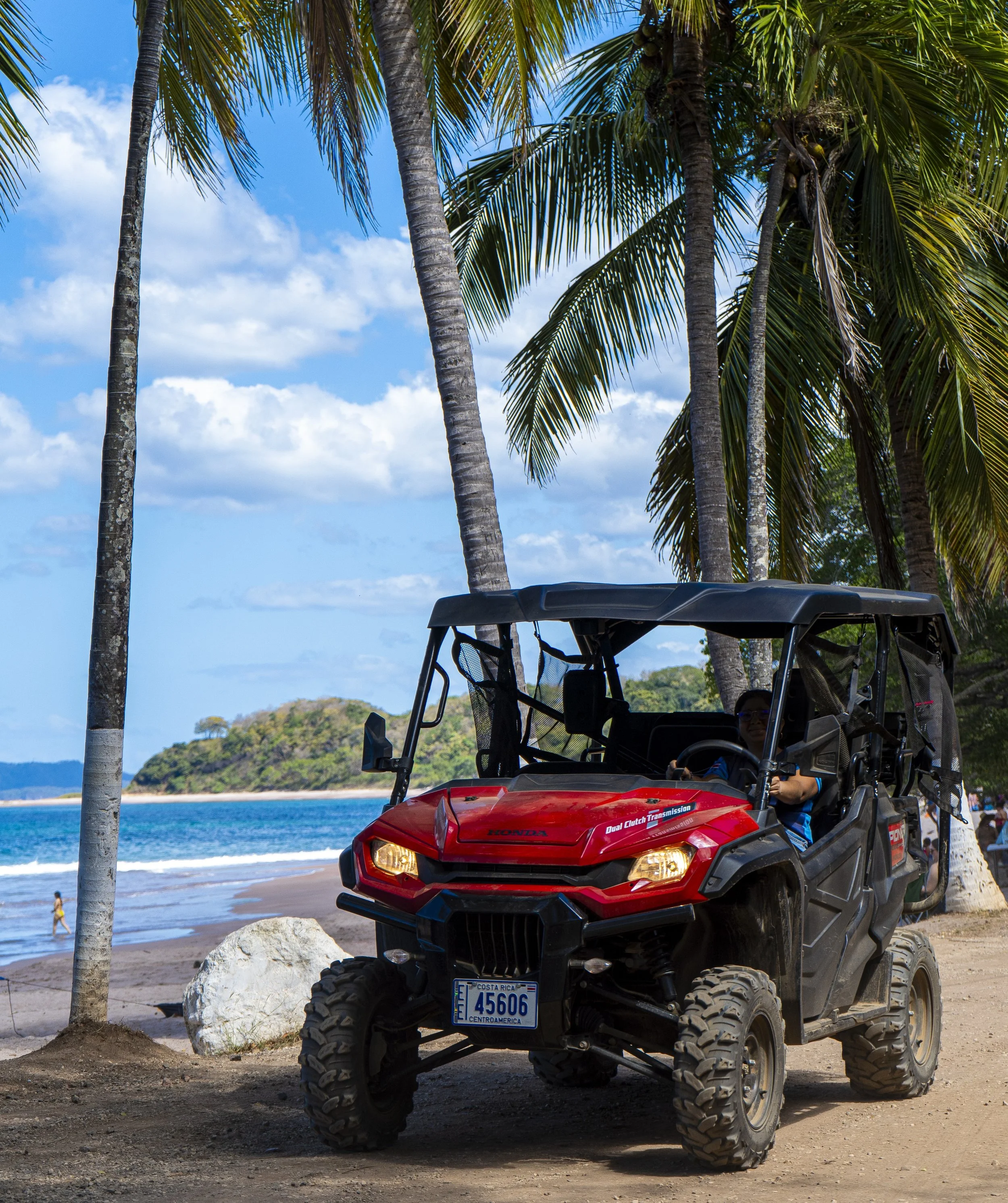 Vehículo todo terreno rojo estacionado en la playa entre palmeras, con mar y cielo azul con nubes en el fondo.