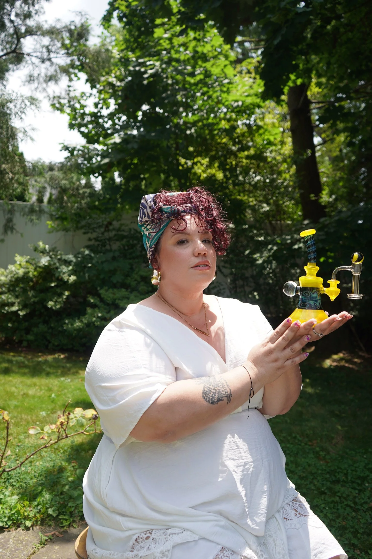 A woman sitting outdoors holding a colorful glass bong, wearing a white dress and a patterned headscarf, with trees and greenery in the background.
