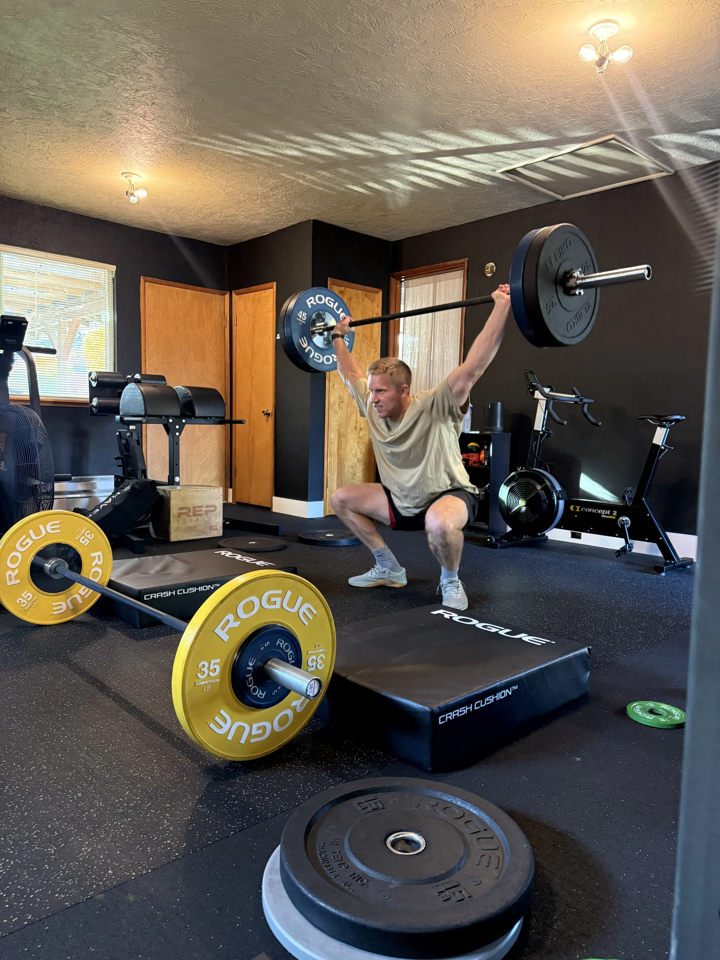 A man is squatting in a gym, lifting a barbell overhead. The gym has weights, a stationary bike, and various workout equipment, with black walls and a window.