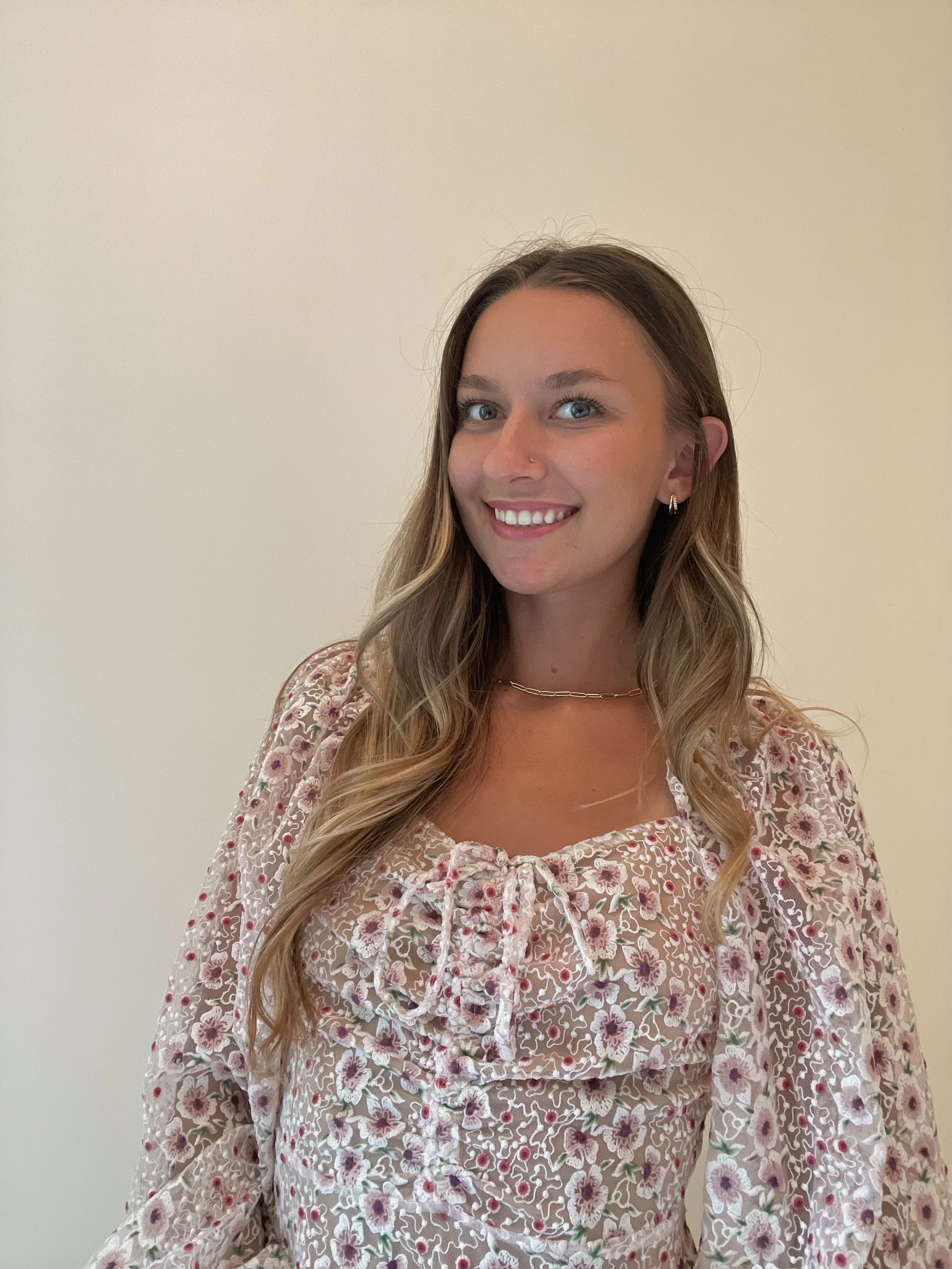 A woman with long wavy hair, blue eyes, and a nose piercing smiling, wearing a floral blouse with ruffles and a thin gold necklace, standing against a plain light-colored wall.