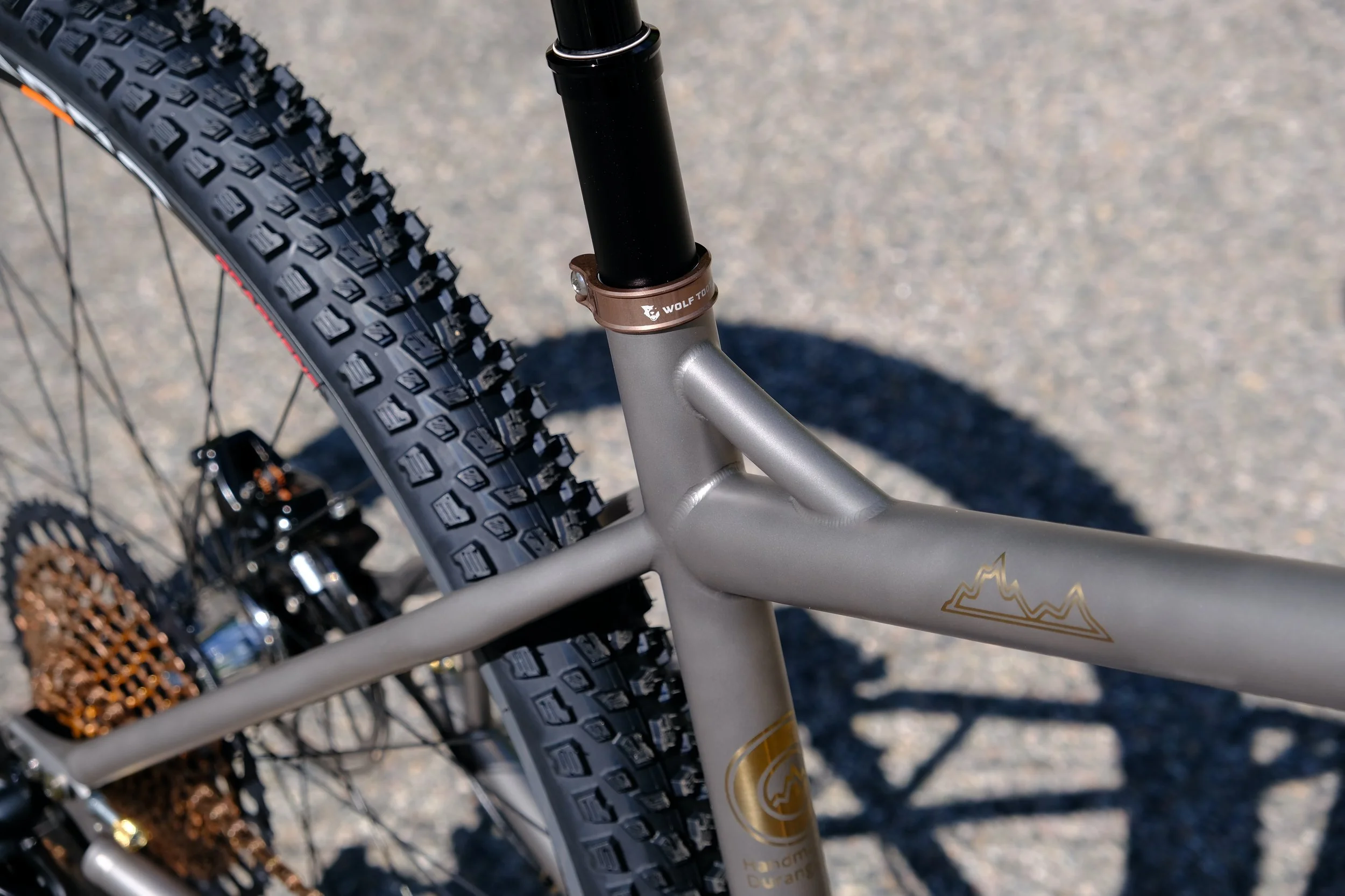 Close-up of a mountain bike's rear wheel, suspension, and frame on a gravel surface.