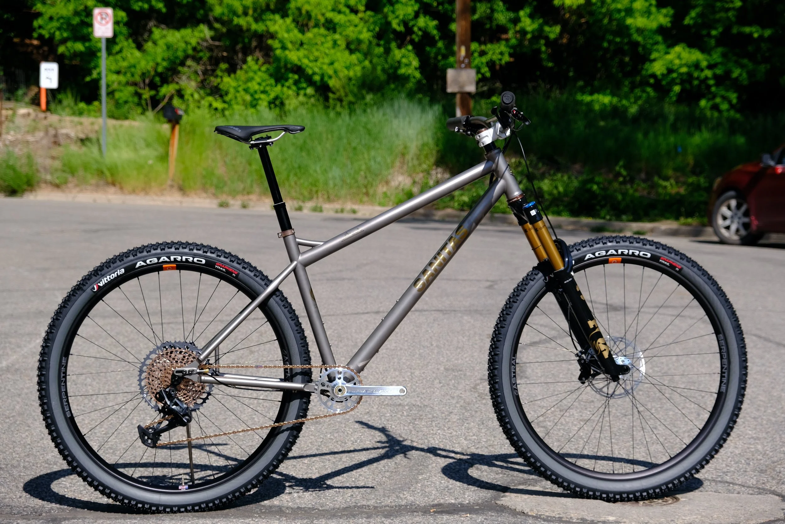 A modern mountain bike with a metallic gray frame, black front suspension fork, and gold anodized accents, parked on an asphalt surface with green trees and a car in the background.