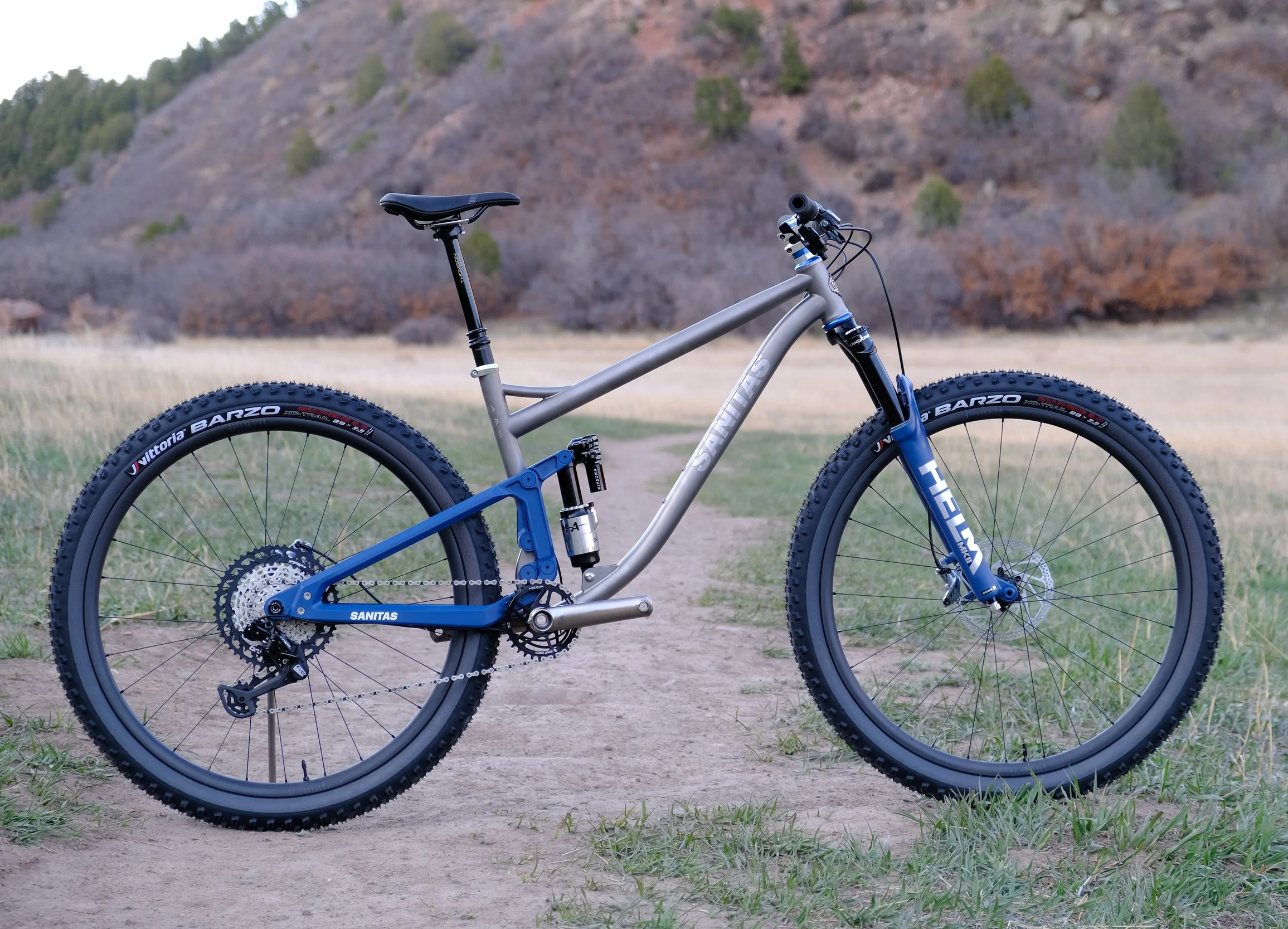 A mountain bike with a silver frame and blue components, sitting on a dirt trail with grassy sides, in a landscape with dry grass and a rocky hillside in the background.