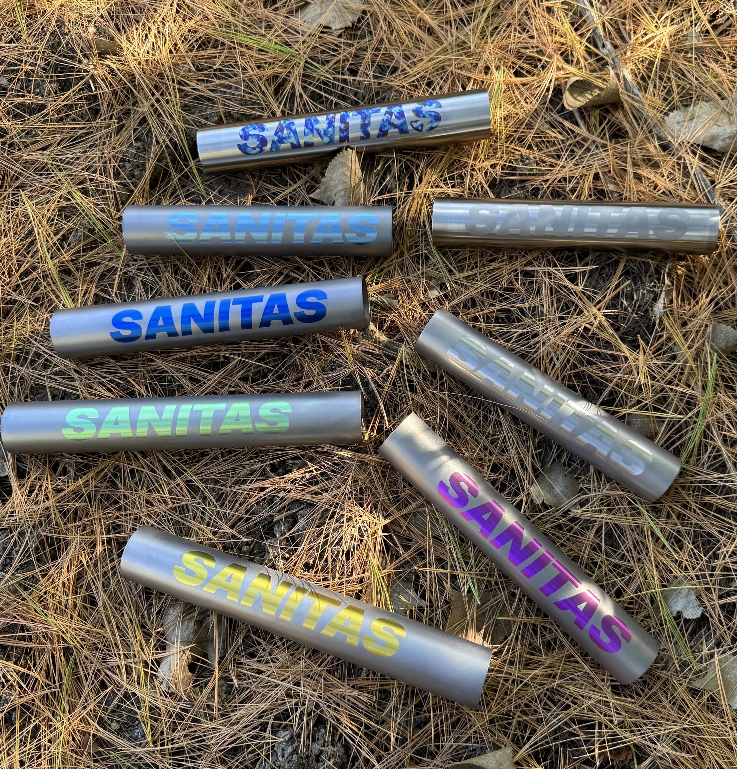 Multiple metallic tubes with the word 'SANITAS' in various anodized colors, placed on a bed of dry pine needles.
