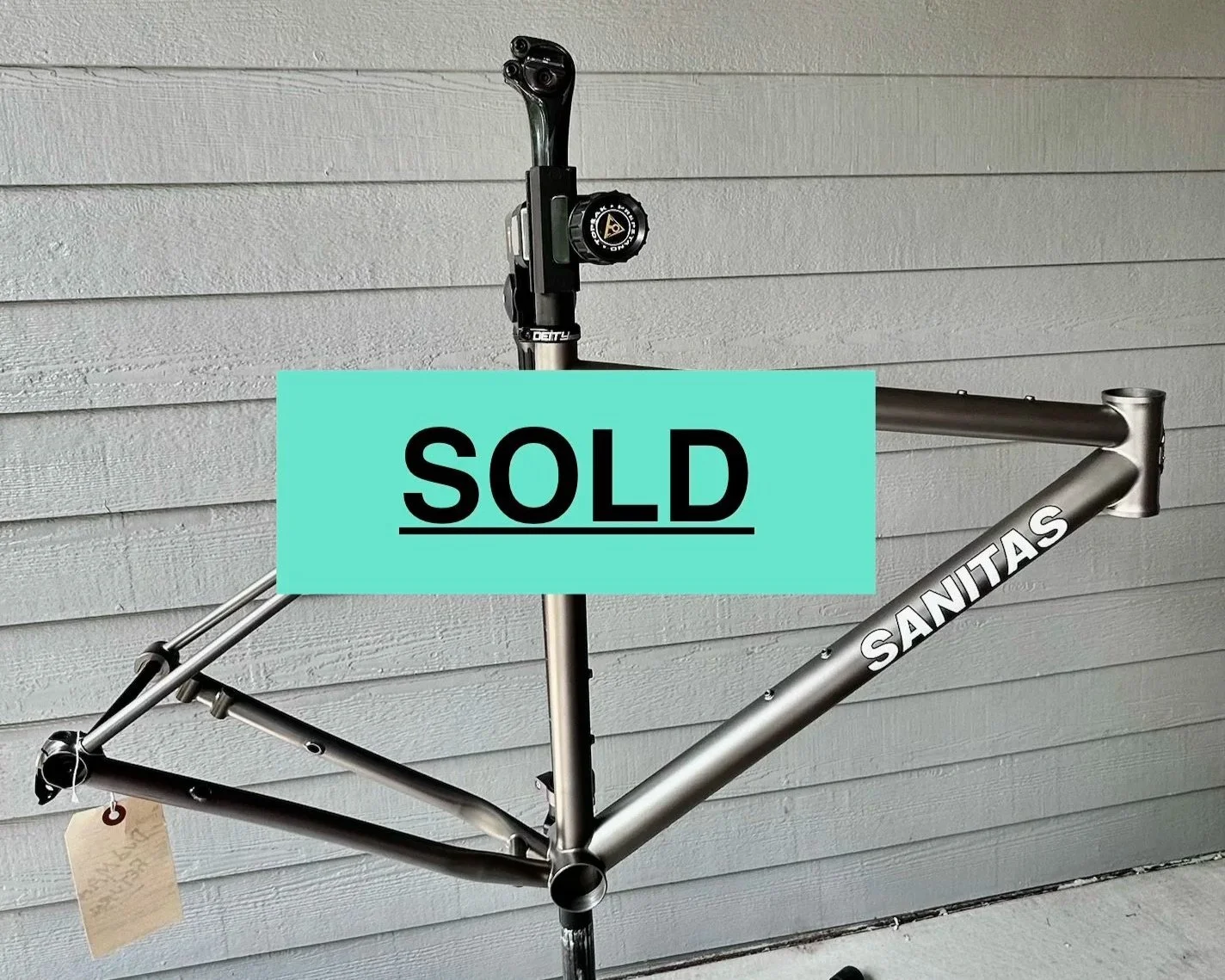 A bicycle frame labeled 'Sanitas' mounted on a stand outdoors against a gray wooden wall.