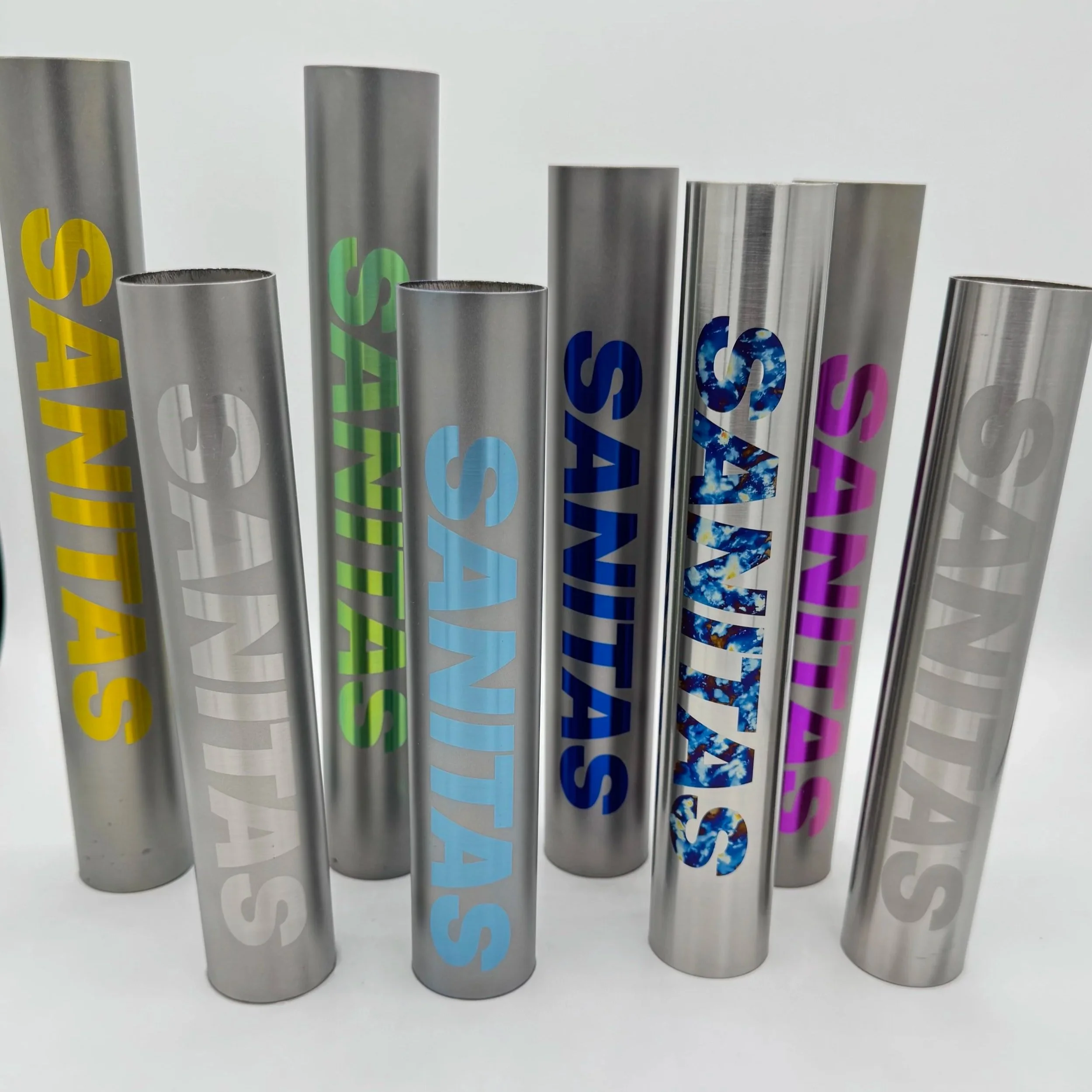 Various metallic tumblers with colorful anodized SANITAS logos in yellow, white, green, light blue, dark blue, galaxy blue, purple, and silver, arranged on a white background.