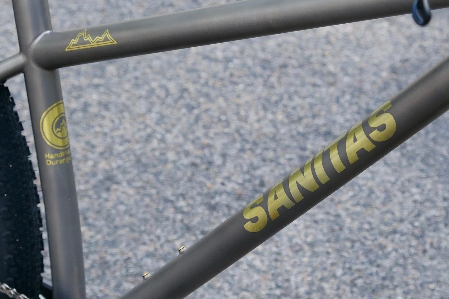 Close-up of a gray mountain bike frame with yellow decals, including the anodized brand name 'SANITAS' and a mountain logo with the words 'Handmade Durango' near the rear wheel.