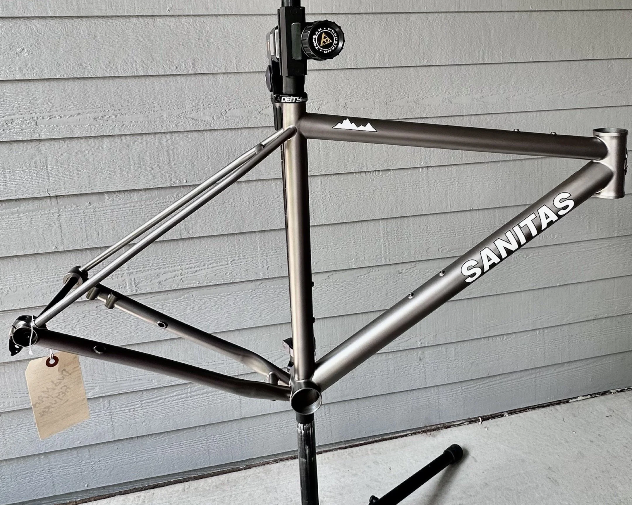 A bicycle frame labeled 'Sanitas' mounted on a stand outdoors against a gray wooden wall.