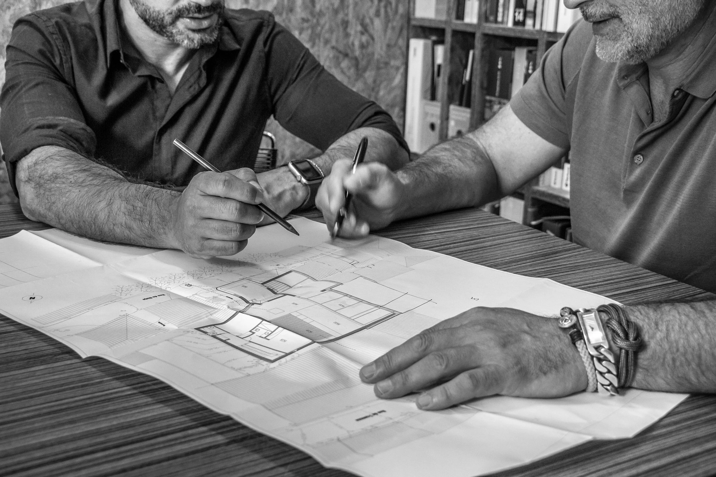 Duas pessoas analisando um projeto de arquitetura ou planta baixa sobre uma mesa, usando canetas para apontar detalhes.