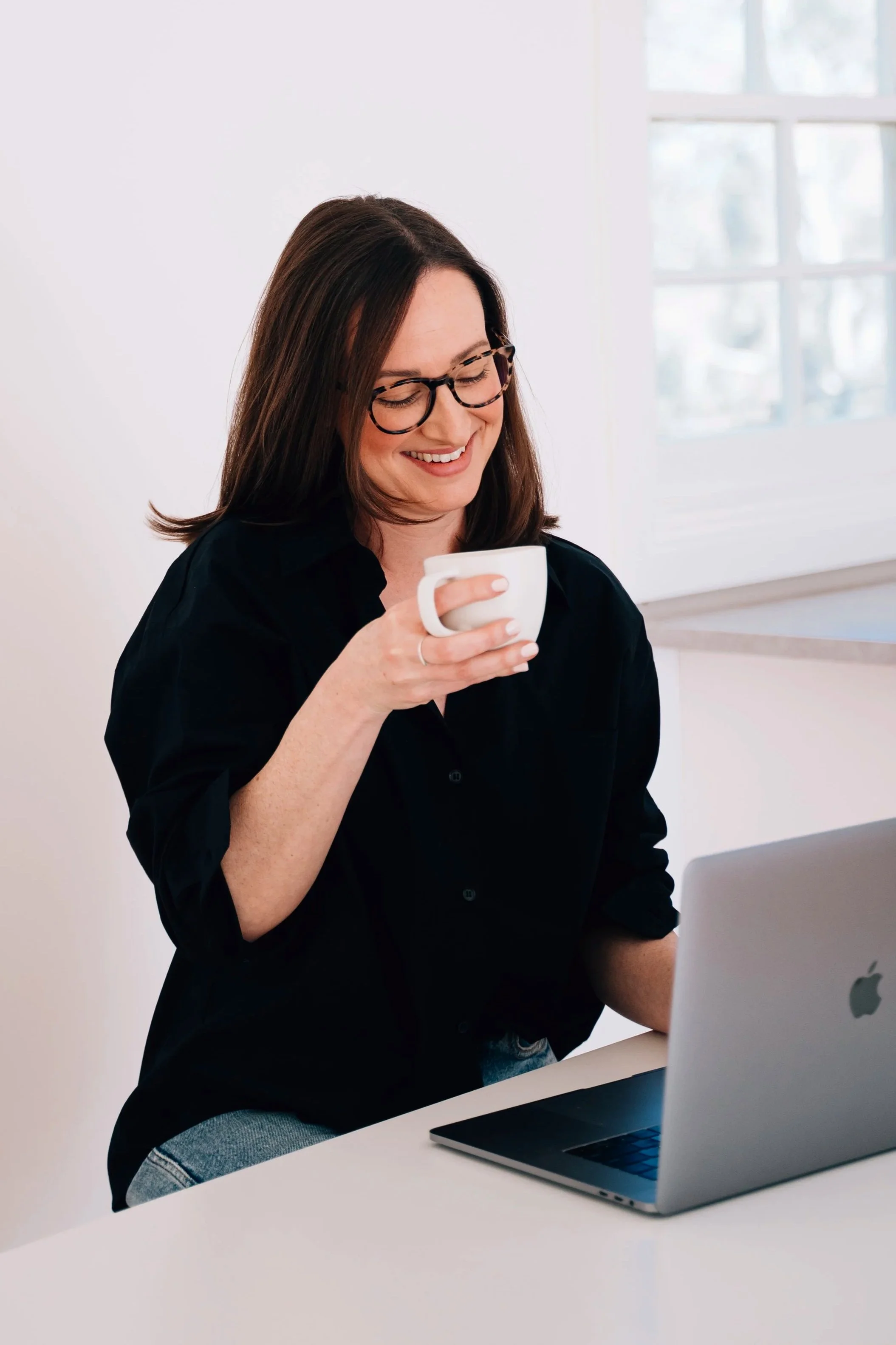 A woman with dark hair, glasses, and a black shirt sitting at a white desk, smiling, holding a white mug, with a laptop in front of her and a window in the background.