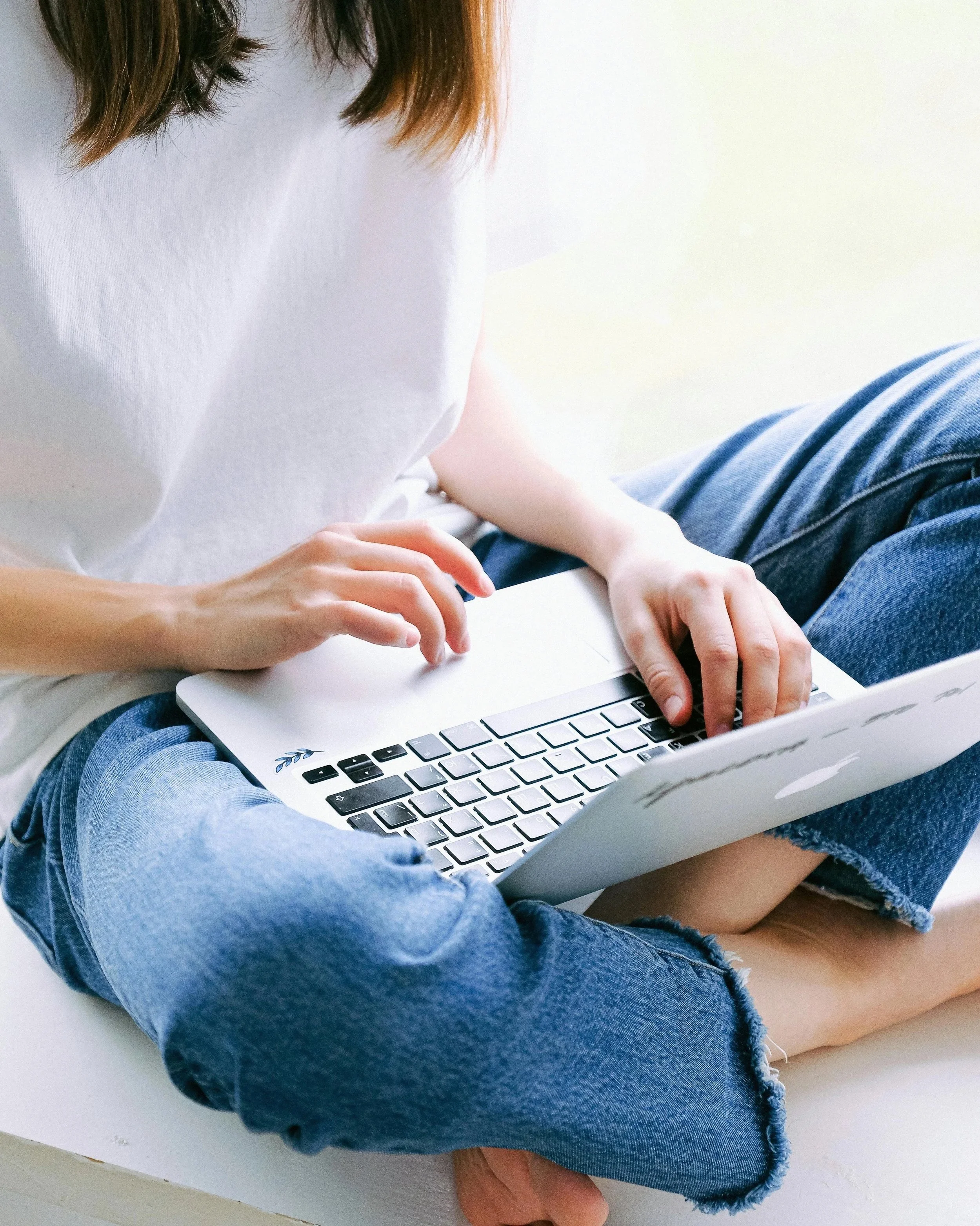Person sitting cross-legged, wearing a white t-shirt and blue jeans, working on a silver laptop.