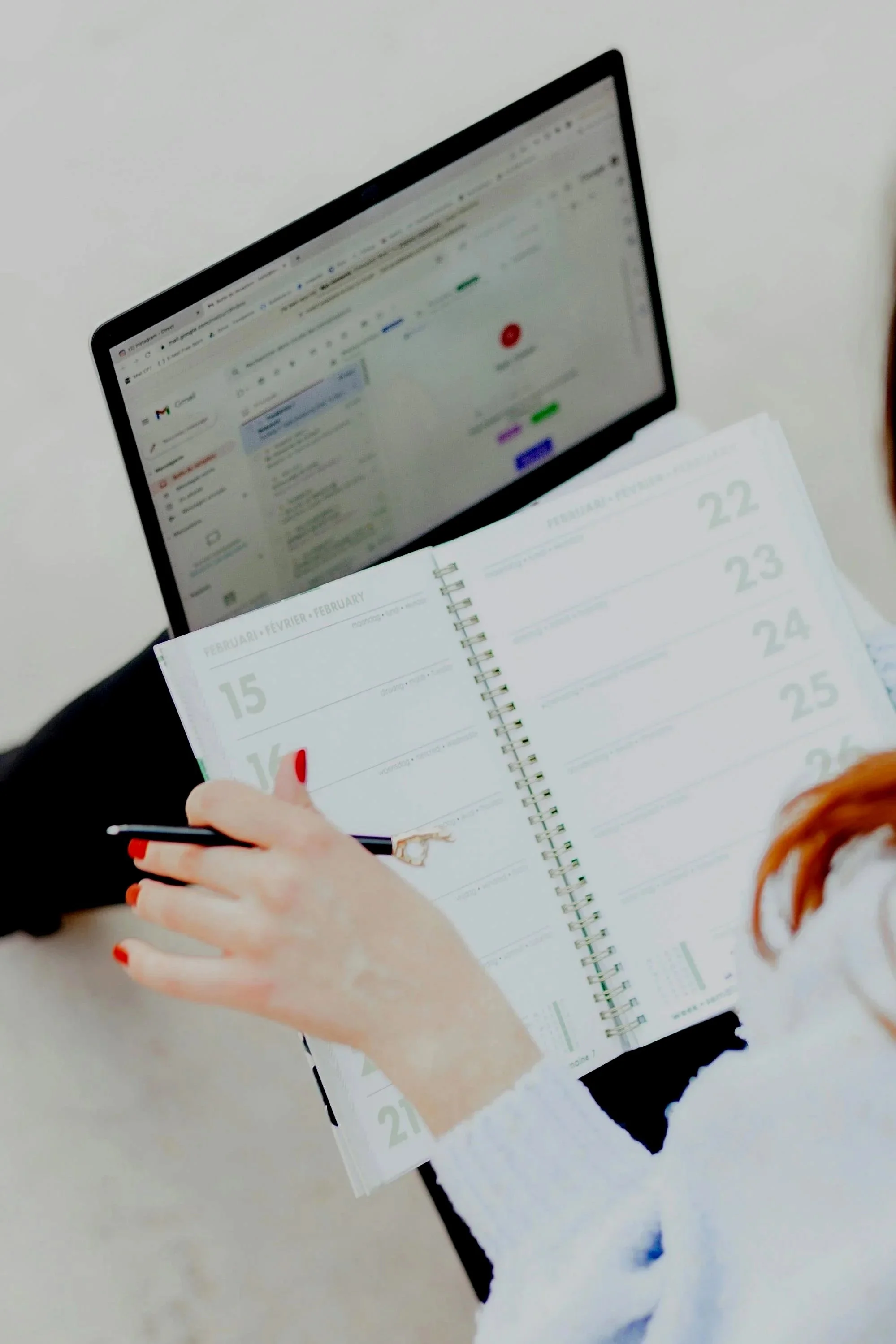A person with red hair is holding a planner and a pen, looking at a laptop screen with an email client open.