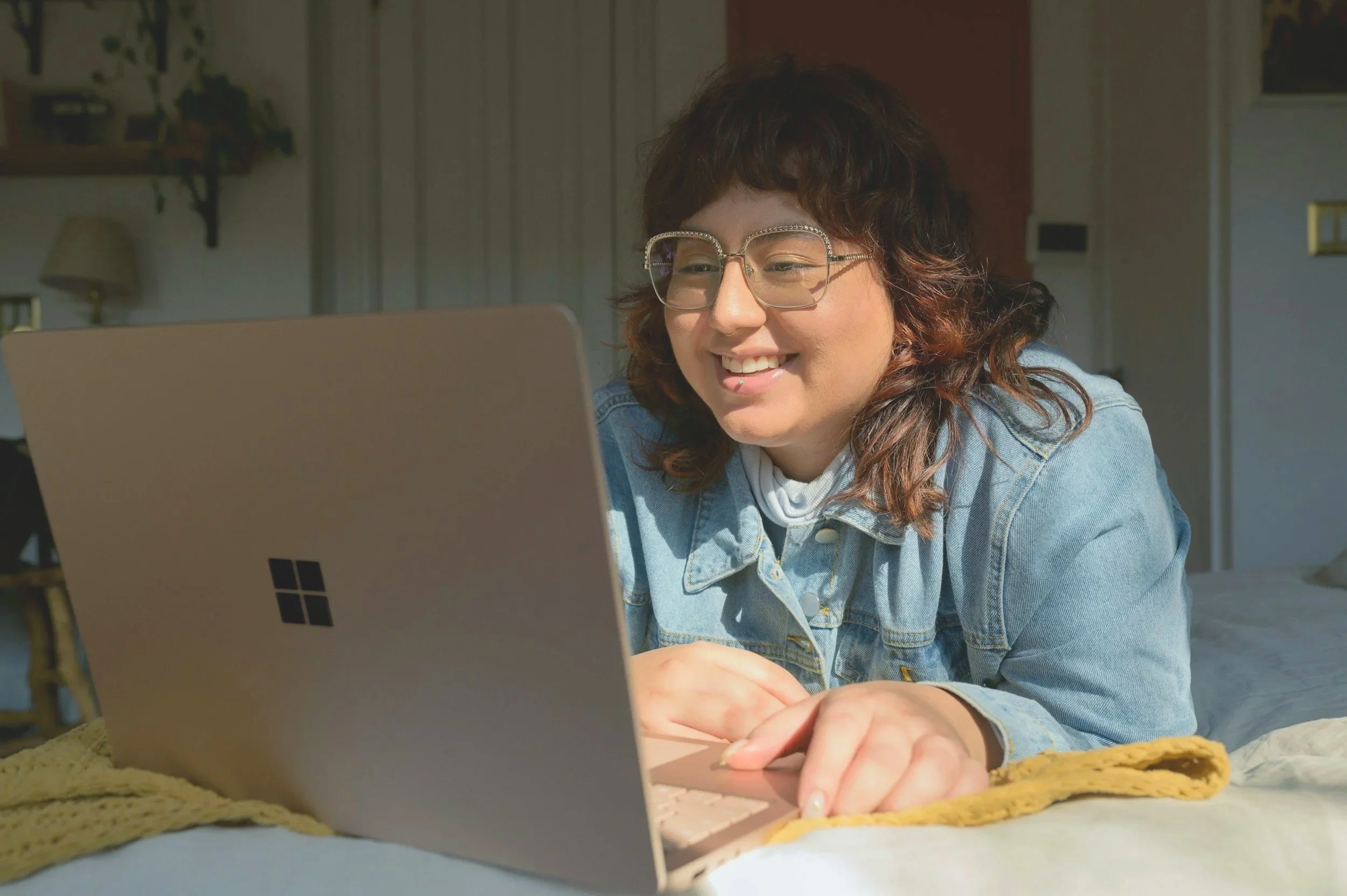 A woman with brown curly hair, glasses, and a denim jacket smiling while using a pink laptop on a bed.