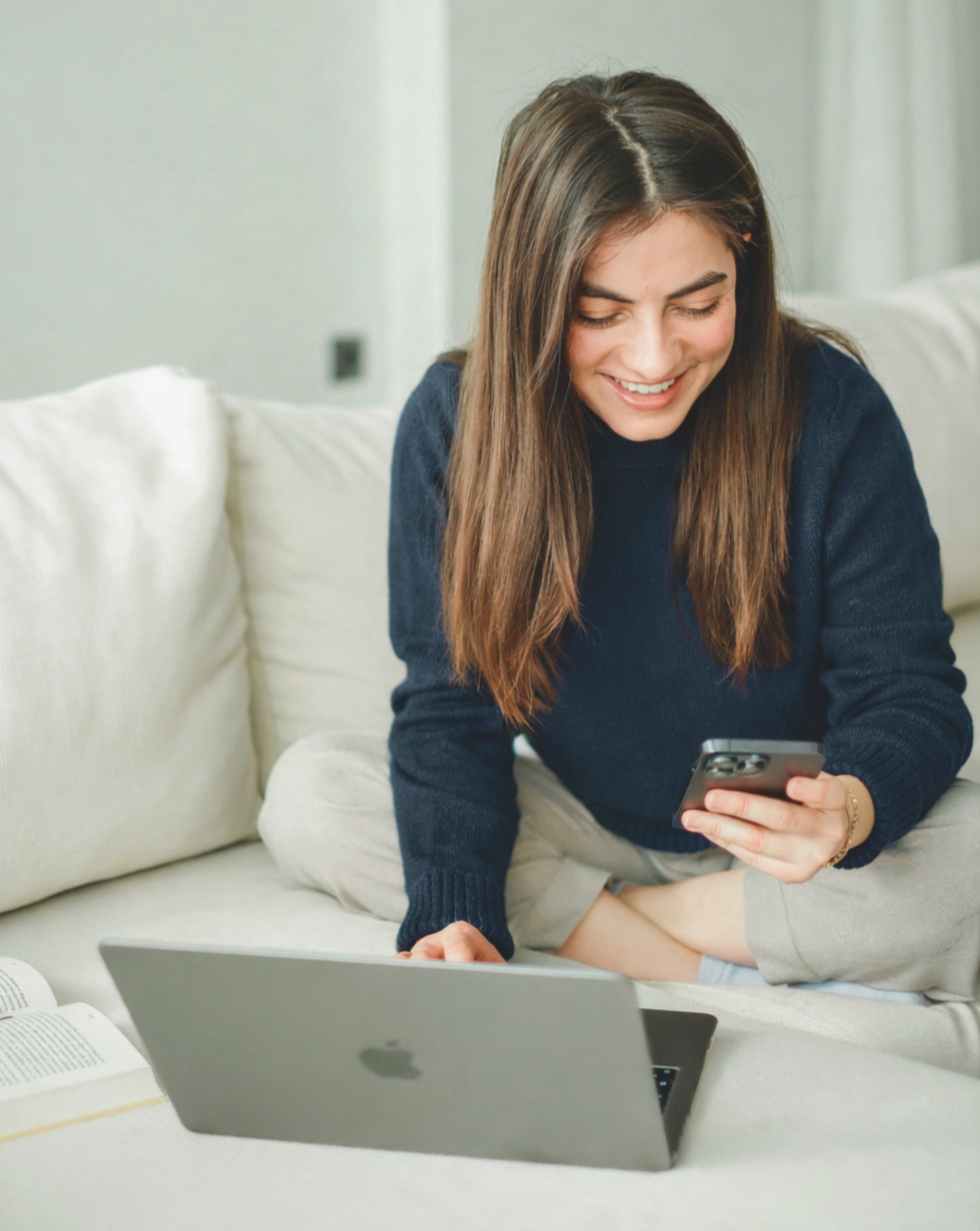 A young woman sitting on a couch, smiling while looking at her phone, surrounded by a laptop, open book, and a pillow.