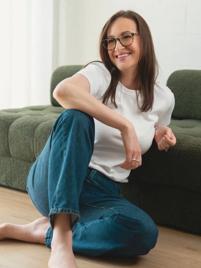 A woman with glasses, long brown hair, wearing a white t-shirt and jeans, sitting on the floor next to a green couch, smiling.