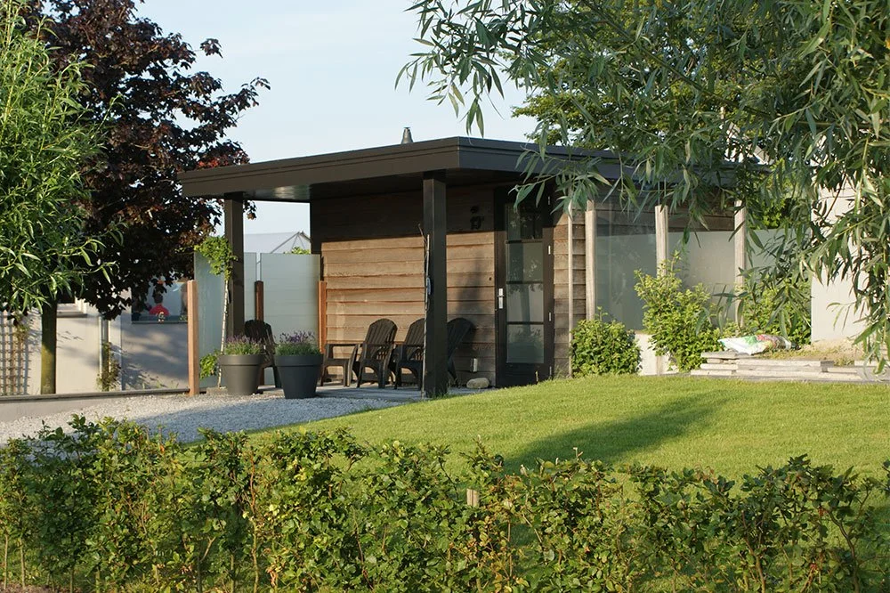 Kleine moderne houten tuin- of tuinkantoor met een overkapping, omringd door een groene tuin met bloemen en bomen.
