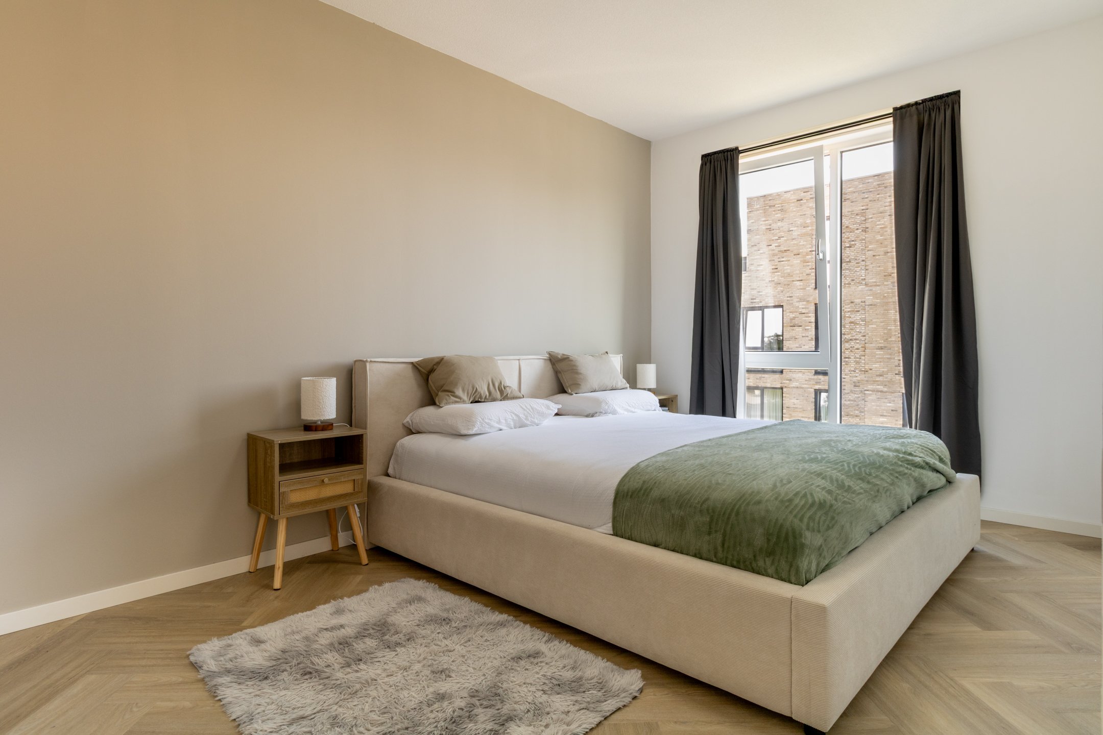 Slaapkamer met beige bed, groene deken, nachtkasten, lampen, groot raam met donkere gordijnen en houten vloer.